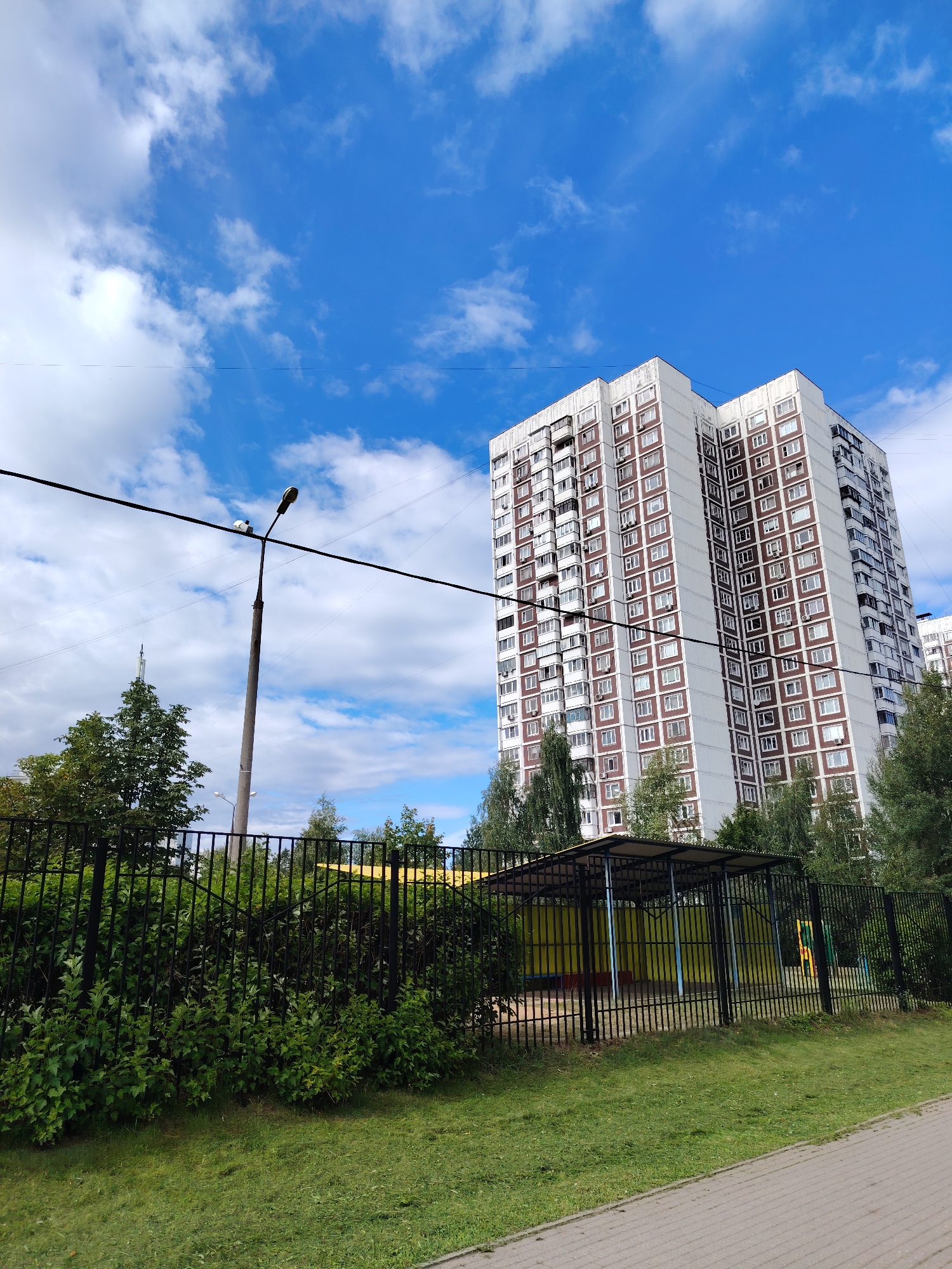 A tall, multi-story residential building with a white and reddish-brown facade dominates the background of this image, set against a bright blue sky dotted with white clouds. In the foreground, a black metal fence encloses a green grassy area with dense bushes and trees. A street lamp with a camera attached to it stands to the left of the building, connected by electrical wires that stretch across the frame. Beyond the fence, a covered structure with a yellow interior and a black metal frame appears to be part of a playground or recreational area. The scene is located in the city of Zelenograd, Russia.