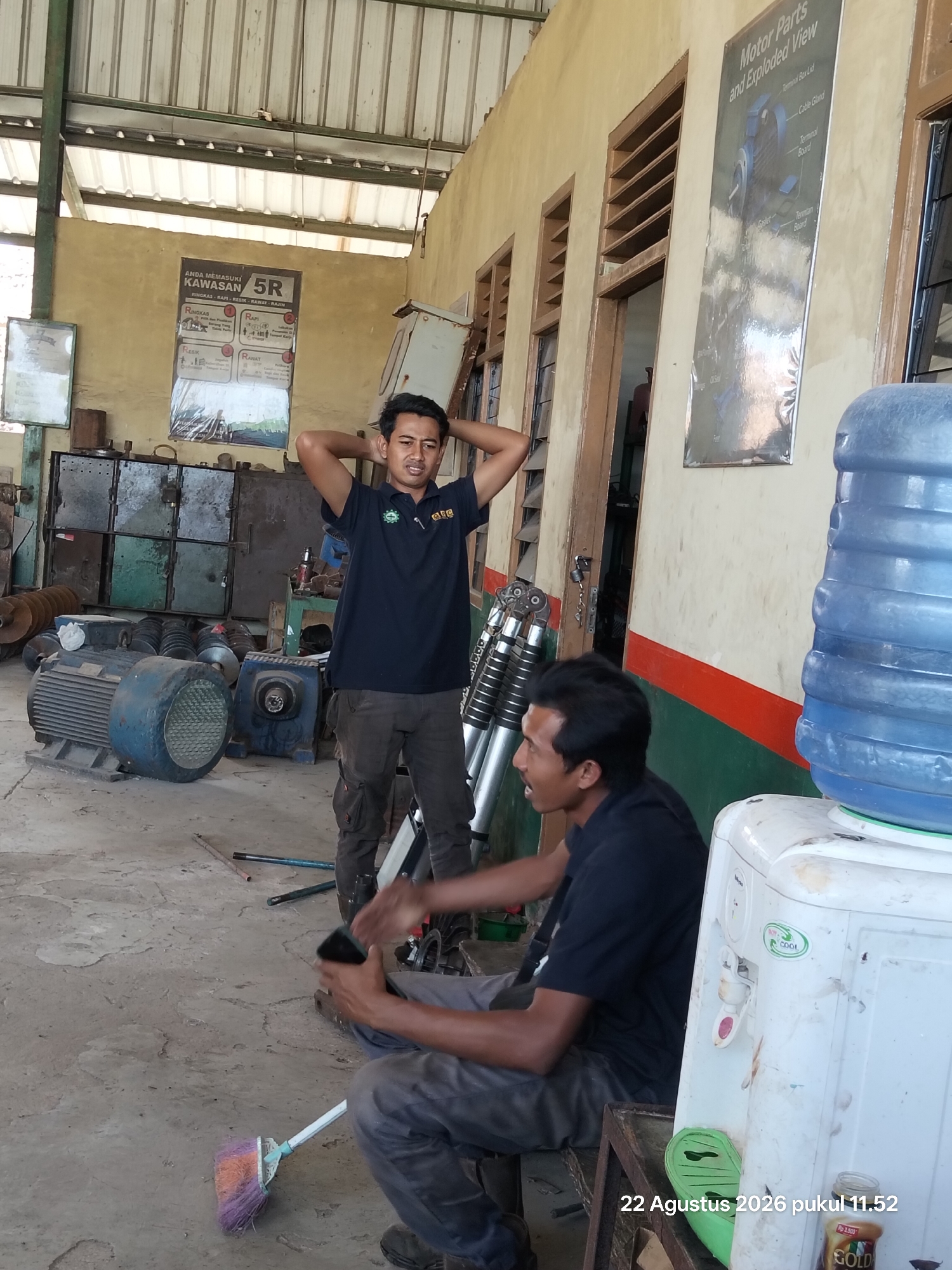 Two men are present in a workshop environment. The man on the left is standing with his hands behind his head, wearing a dark blue t-shirt with a logo and dark grey cargo pants. The man on the right is seated on a low bench, holding a black smartphone in his right hand, and wearing a dark blue t-shirt and dark grey pants. In the background, there are various industrial equipment, including large blue motors, metal cabinets, and a water dispenser. A broom with multi-colored bristles is on the floor in the foreground. A poster detailing the "5R" principles (Ringkas, Rapi, Resik, Rawat, Rajin) is on the wall to the left, and another poster with a diagram of "Motor Parts and Exploded View" is on the wall to the right. The floor is concrete, and the walls are a light yellow color. The roof appears to be corrugated metal. The timestamp "22 Agustus 2026 pukul 11.52" is visible in the bottom right corner, suggesting the image was taken in Indonesia.