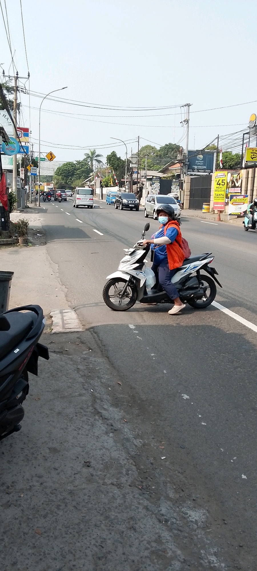 A person wearing a blue shirt, orange vest, and helmet rides a white motorcycle on a paved road in Depok, Indonesia. The person is also wearing a blue face mask and dark pants. The motorcycle has a red backpack on the back. The road has white lane markings and is lined with trees and buildings. Other vehicles, including cars and motorcycles, are visible in the background. The sky is clear and overcast. The image appears to be taken during the daytime.