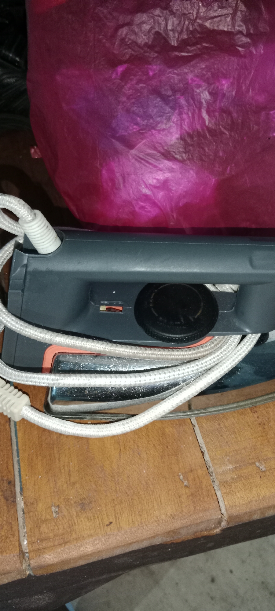 In Subang, Indonesia, an iron rests on a wooden surface. The iron is gray with a white cord coiled around it. A pink plastic bag is visible in the background. The lighting is dim, creating a shadowy atmosphere. The image focuses on the iron, highlighting its metallic base and plastic casing.