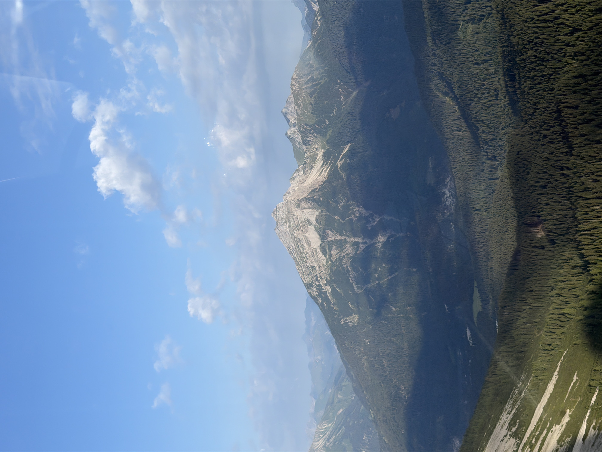 The image shows a vast mountainous landscape under a bright blue sky with scattered white clouds. In the foreground, a steep, rocky slope is visible, transitioning into a dense forest of evergreen trees that covers the lower slopes of the mountains. The midground is dominated by a large, rugged mountain with visible rocky outcrops and vegetation. Further in the distance, more mountain ranges fade into a hazy blue. The scene is bathed in sunlight, casting shadows across the forested valleys and highlighting the textures of the mountains. The location is identified as Grainau, Germany.