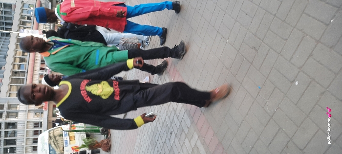 The image depicts an urban street scene in Nairobi, Kenya, with several individuals walking on a paved sidewalk. In the foreground, a young male of African descent walks towards the left side of the frame. He wears a long-sleeved black shirt with yellow cuffs and collar trim, emblazoned with a green cartoon character and the red text "NEVER ROUGH" above it and yellow text "ALWAYS NICE!" below. He wears black trousers and light brown shoes, holding a white smartphone in his right hand.
Behind him and to his right, an adult male of African descent walks in the same direction, wearing a green work-style jumpsuit with blue accents and black shoes. He carries a white and black patterned bag in his left hand. To the far right, another adult male of African descent is partially visible, wearing a red long-sleeved garment with green and white trim, blue trousers, and a blue cap, with black shoes. Additional individuals are present in the blurred background, including a woman in a white head covering.
The ground is paved with grey interlocking rectangular bricks laid in a herringbone pattern. In the background, multi-story concrete buildings with multiple windows line the street, characteristic of a commercial or downtown area. A white passenger van, consistent with a Kenyan matatu, is partially visible on the far left, positioned near a small plant. All visible individuals are in motion, walking along the sidewalk. No direct interactions between individuals are discernible.