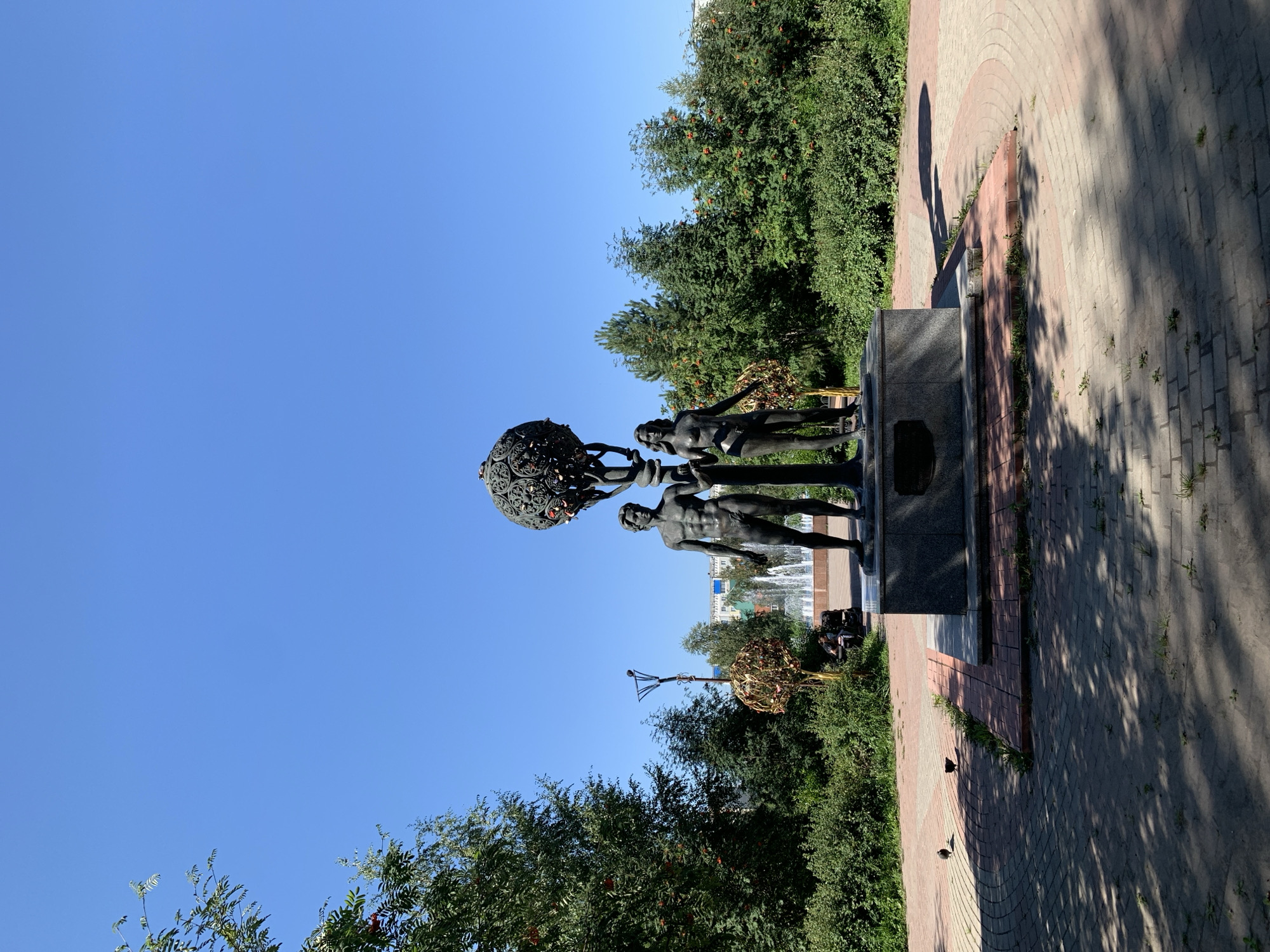 The image shows a bronze sculpture of Adam and Eve in a park in Prokopyevsk, Russia. The sculpture is located on a pedestal in the center of a paved area with trees and bushes in the background. A fountain is visible behind the sculpture. The sky is clear and blue.
