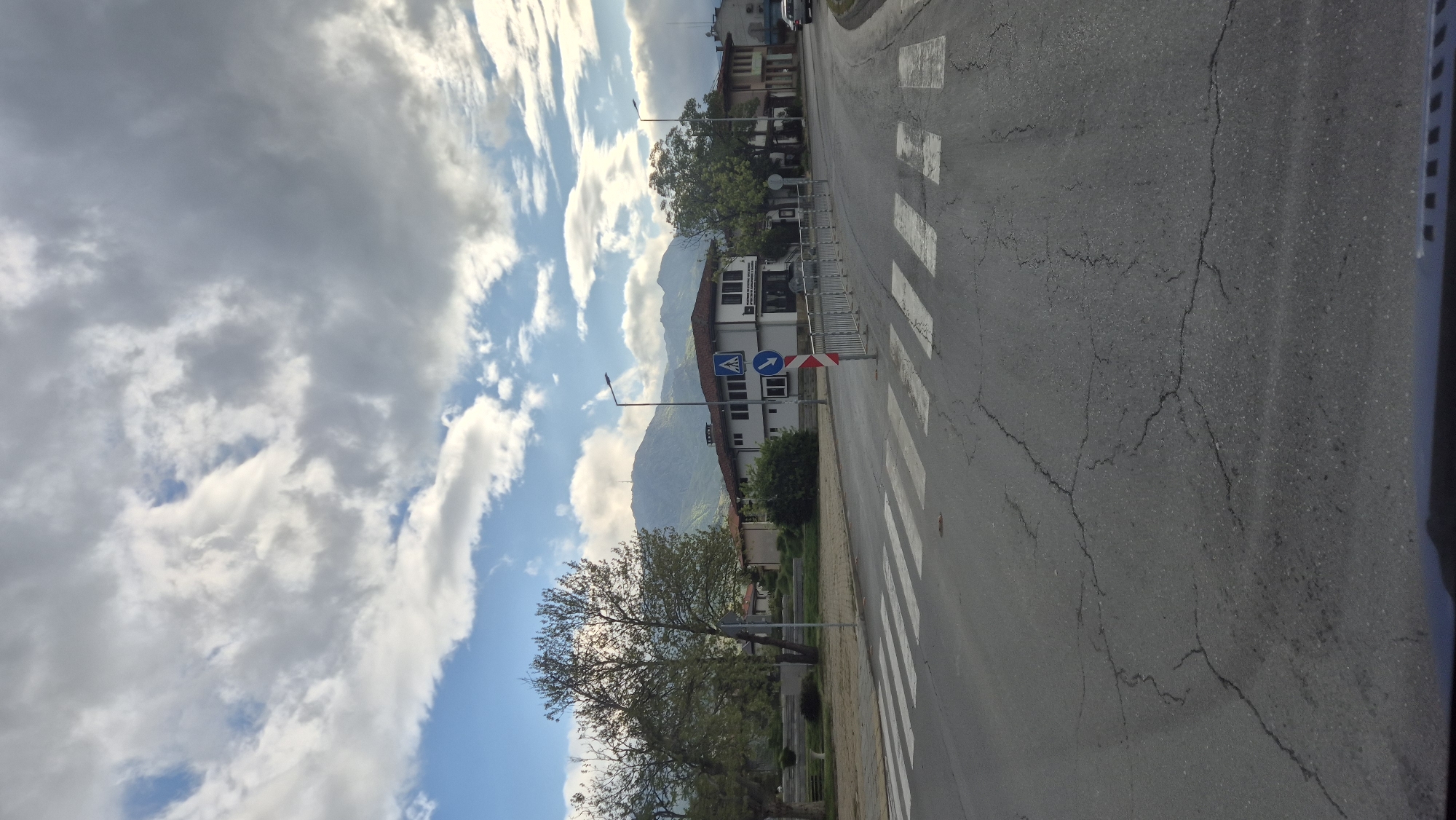The image depicts a street scene in Sliven, Bulgaria. The foreground features an asphalt road with a pedestrian crosswalk, marked by white stripes. The road surface is cracked. A pedestrian crossing sign stands adjacent to the crosswalk, indicating a designated area for pedestrians. To the left of the road, a tree with sparse foliage and a grassy area. In the background, there is a two-story building with a dark roof and a white facade. Behind the building, a mountain rises, partially obscured by clouds. The sky is filled with a mix of blue sky and white clouds.
