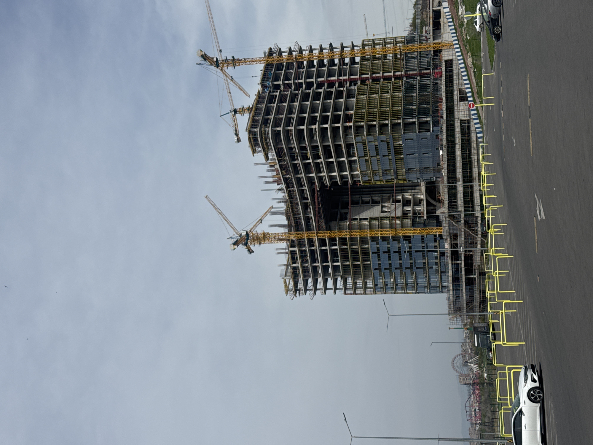 The image captures a tall, unfinished building under construction in Yerevan, Armenia. Several yellow cranes are positioned around the structure. The building appears to be a modern high-rise with an exposed skeletal framework. A wide road borders the construction site, lined with yellow guardrails. Beyond the road, there is a partially obscured view of a Ferris wheel, suggesting an amusement park or recreational area in the vicinity. The sky is overcast. Several cars are parked and driving on the street.
