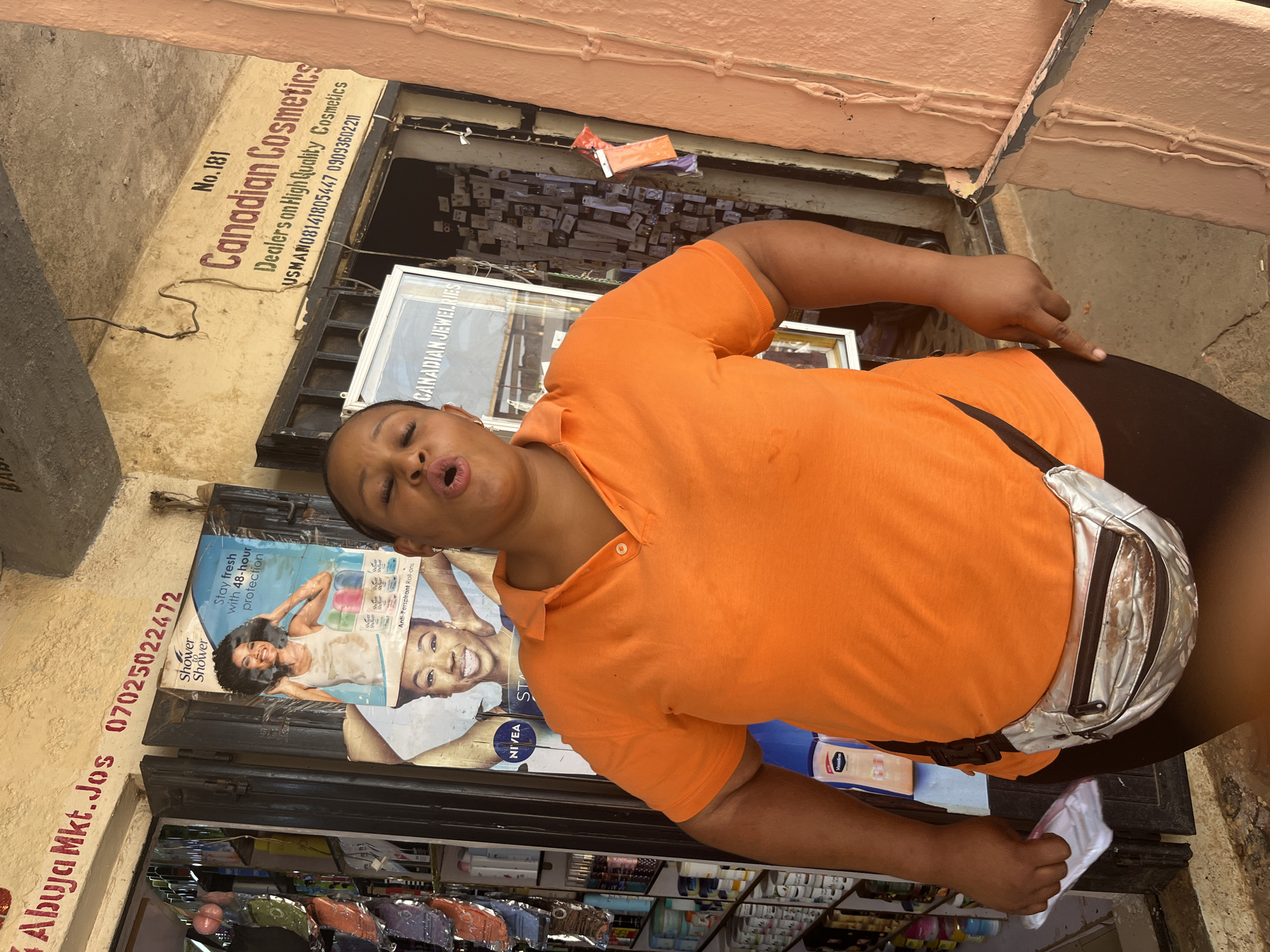 A woman wearing an orange polo shirt and a silver fanny pack stands in front of a storefront in Jos, Nigeria. She is looking directly at the camera with her mouth open. The storefront has a sign that reads "Canadian Cosmetics" and another sign in red paint above that states "Abuja Mkt. Jos 07025022472". Various products are visible on shelves behind the woman. The scene appears to be an outdoor market.