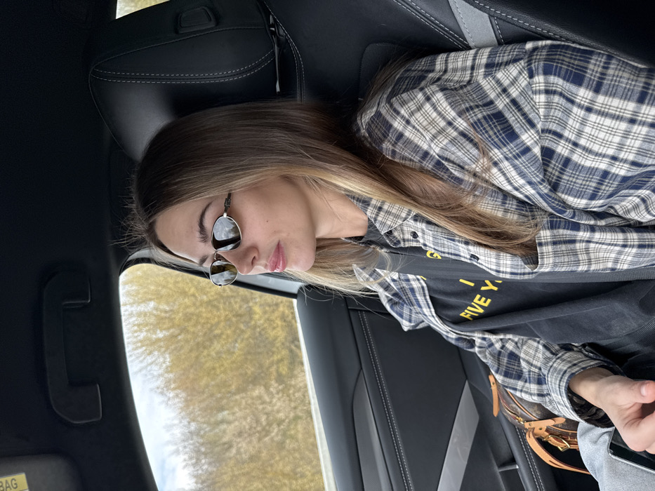 A female individual is seated as a passenger inside a vehicle in Pasohlávky, Czechia. She is positioned facing left, looking out the window. The individual has long, light brown hair with lighter highlights and is wearing small, oval-framed sunglasses with reflective lenses. Her attire consists of a dark-colored t-shirt with yellow text, partially visible as "FIVE Y...", layered under an open blue and white plaid long-sleeve shirt. A black seatbelt is fastened across her chest and lap. To her right, a small brown bag with a distinctive pattern is visible on the seat. The vehicle's interior features dark upholstery, including a black seat with a visible headrest stitching pattern and a dark door panel. Outside the window, a landscape of yellowish-brown vegetation, likely trees or bushes, is visible under a light blue sky with some cloud cover. A grey barrier or guardrail runs horizontally below the vegetation.