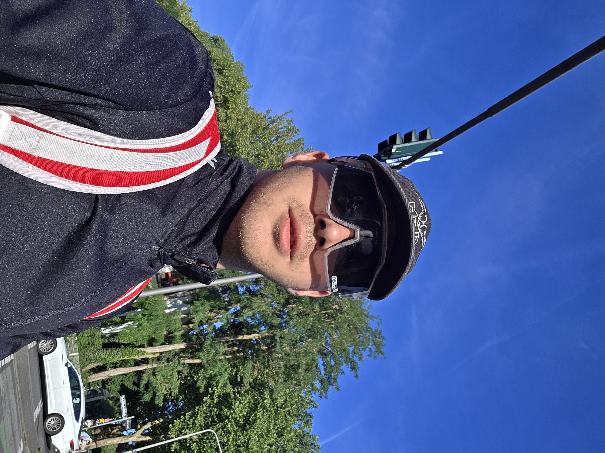 A medium shot captures a man wearing sunglasses and a baseball cap, looking directly at the camera. He is outdoors, with a clear blue sky above and lush green trees in the background. A traffic light is visible in the upper left quadrant of the image, and a white car is driving on a street to the right. The man is wearing a black jacket with red and white stripes on the shoulders. He appears to be in a city, possibly preparing for or returning from an outdoor activity, such as cycling, given his attire. The lighting suggests it is daytime.