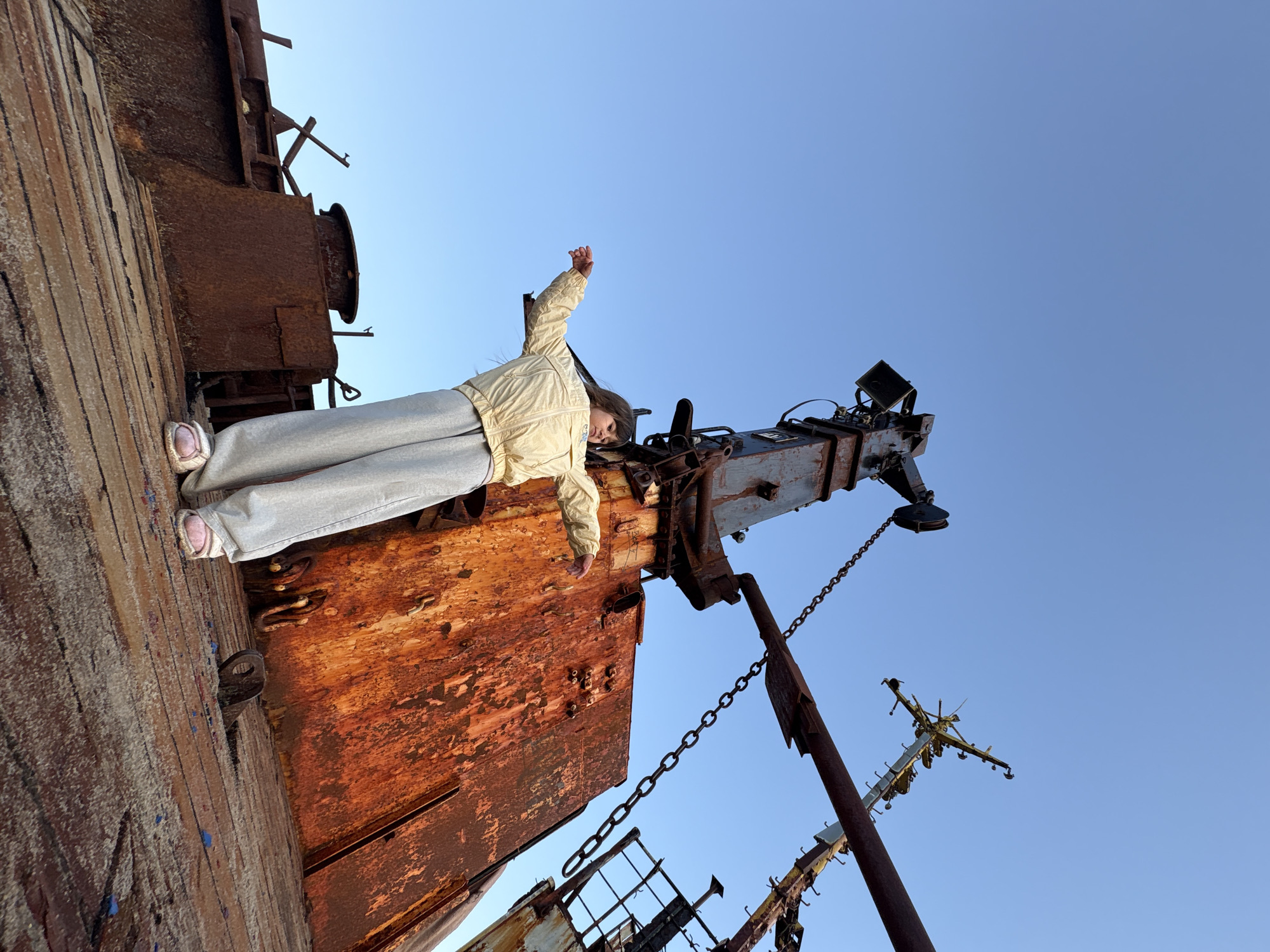 A young girl stands on the deck of a rusty shipwreck in Teriberka, Russia. She is wearing a light yellow jacket and light grey sweatpants. Her arms are spread wide as if embracing the vastness of the sky. The shipwreck, heavily weathered and covered in rust, dominates the foreground and middle ground. In the background, a clear blue sky is visible. The scene suggests a sense of exploration and wonder amidst the decaying remnants of maritime history.