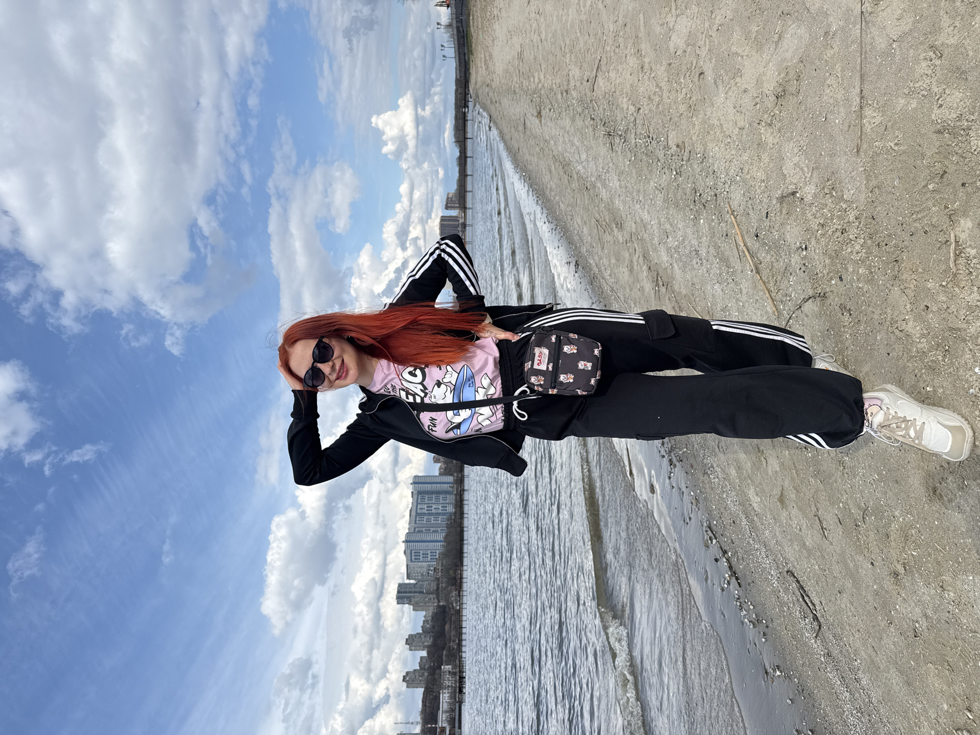 A woman with red hair and sunglasses is standing on a sandy beach in Rostov-on-Don, Russia. She is wearing a pink t-shirt with cartoon characters, a black track jacket with white stripes, black track pants with white stripes, and white sneakers. She has a small black bag with white cartoon characters slung over her shoulder. In the background is a body of water, likely the Don River, with the city skyline visible on the opposite bank. The sky is partly cloudy with blue patches. The photograph appears to be taken at an angle, possibly due to the position of the person taking the picture or to make the picture more interesting.