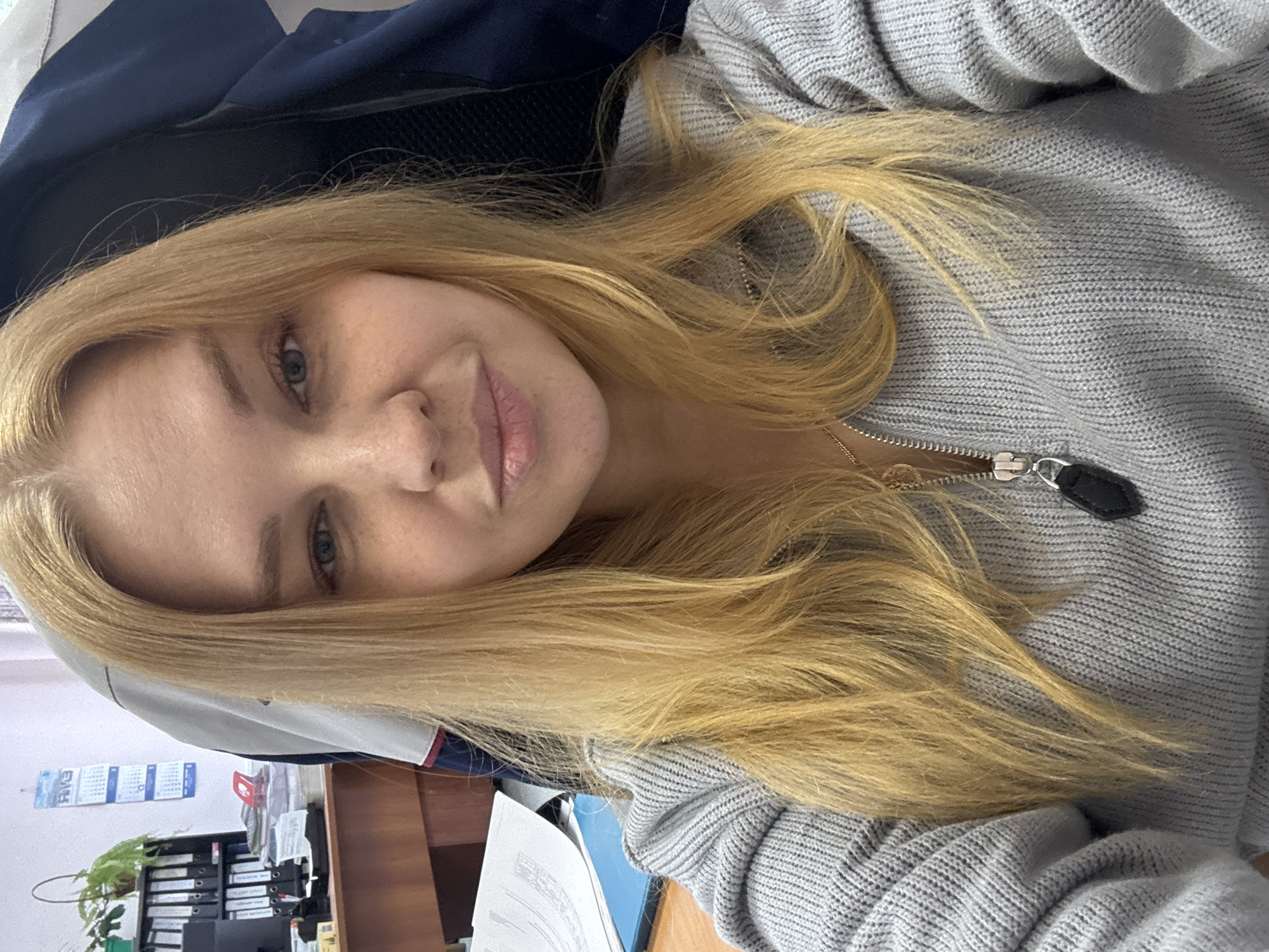 A fair-skinned woman with long, blonde hair is seated in a chair with a dark blue back. She wears a light gray, knitted sweater with a metal zipper and a small black pendant. The setting appears to be an office environment, with a wooden desk visible in the background, cluttered with files, a calendar on the wall, and a plant. She is in Rusanovo, Russia.