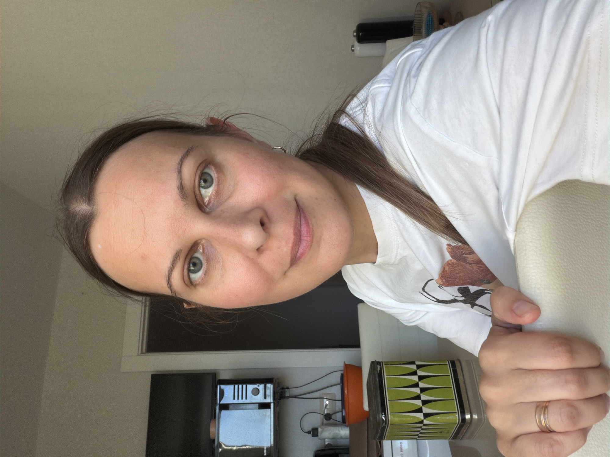 A woman, appearing to be in her 30s to early 40s, is depicted in a chest-up, eye-level shot within an indoor residence in Izhevsk, Russia. She has light-colored eyes and dark blonde hair, parted in the middle and pulled back. A small silver hoop earring is visible in her right ear. She wears a white short-sleeved t-shirt with an abstract brown and black graphic design on the front. Her left hand, adorned with a gold-toned ring on the ring finger, rests on the light-colored, upholstered back of a chair. The woman looks directly at the camera with a slight smile. The background to the left features a wall-mounted black Samsung microwave oven above a partially visible television screen, with wires and an orange bowl on a surface below. A distinctive rectangular metal tin with a green, black, and white geometric pattern is situated on a table in the left foreground. To the right, against a light-colored wall, two cylindrical dispensers, one white and one black, are visible on a surface.