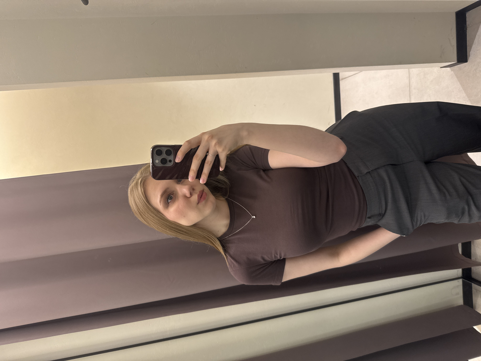A young, fair-skinned woman with straight blonde hair and blue eyes is captured in a mirror selfie within an indoor setting in Perm, Russia. She is wearing a fitted, short-sleeved dark brown top, dark grey tailored trousers with her right hand inserted into a pocket, and a delicate silver necklace featuring a small, distinctive pendant. Her left hand holds a dark-colored smartphone, identified as an iPhone Pro model due to its triple-lens camera array, positioned to take the photograph. A ring is visible on her left ring finger. The reflection in the mirror reveals a changing room or fitting room environment, characterized by a large, brownish-purple curtain to the left and a light cream-colored wall to the right. A dark baseboard runs along the bottom of the wall, and the floor appears to be light-colored tile. The woman displays a slightly pouty facial expression, directing her gaze towards the phone/mirror.