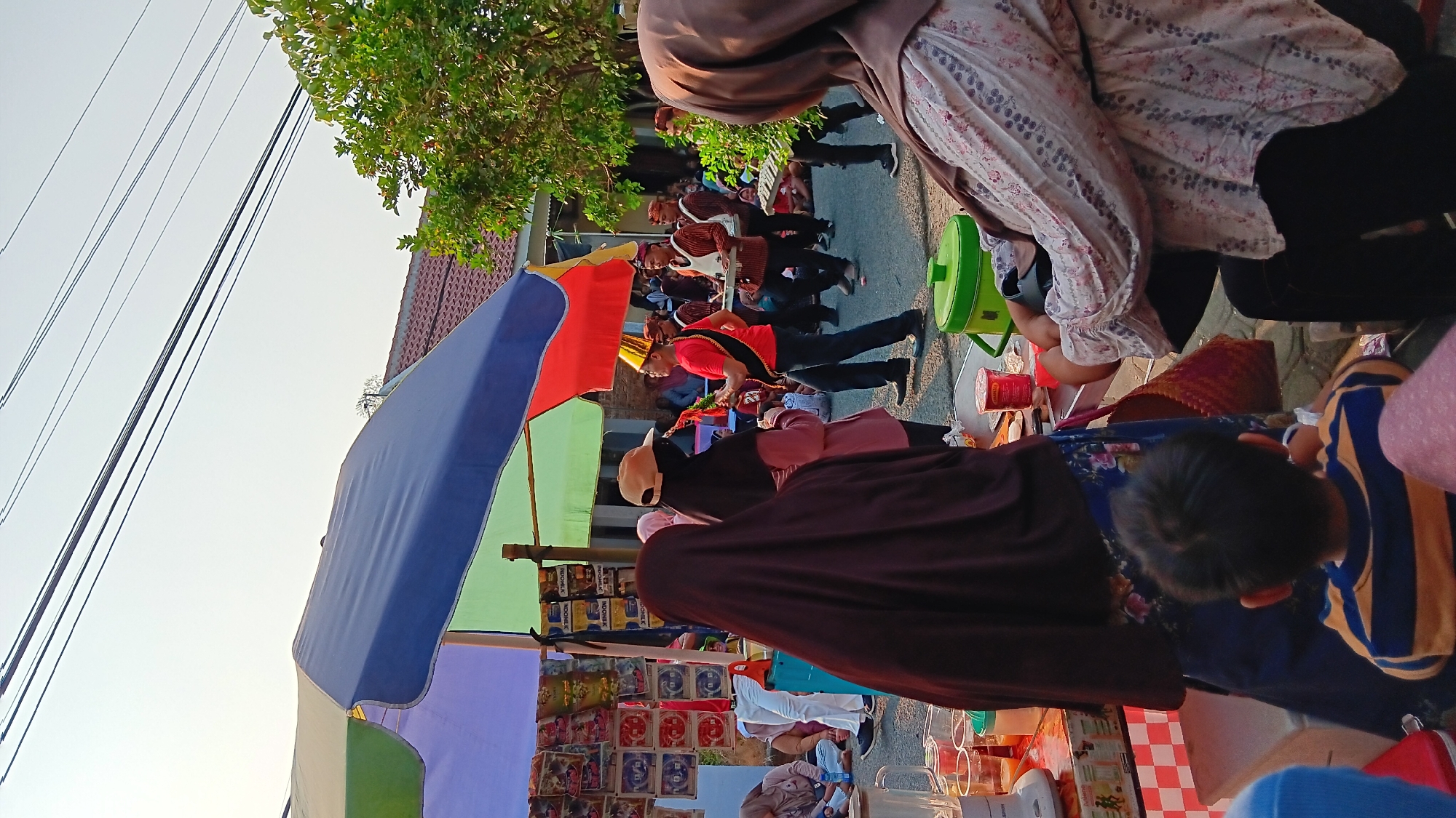 In Cerme, Indonesia, a street scene unfolds beneath a colorful canopy, suggesting a festive or market event. A group of men, dressed in traditional attire including red shirts, black sashes, and headwear, are performing with musical instruments, likely part of a procession or parade. One man in the foreground wears a gold crown-like headdress and plays a green instrument, possibly a xylophone or glockenspiel. Another man carries a similar instrument on a stand. Several spectators are visible, including women wearing hijabs and a young boy looking towards the performers. A stall with various items displayed, possibly for sale, is set up under the canopy. The environment appears to be a street with houses and trees in the background, bathed in the warm light of what seems to be late afternoon or early evening. The ground is paved, and power lines are visible overhead, indicating an urban or semi-urban setting. The overall atmosphere suggests a lively community gathering.