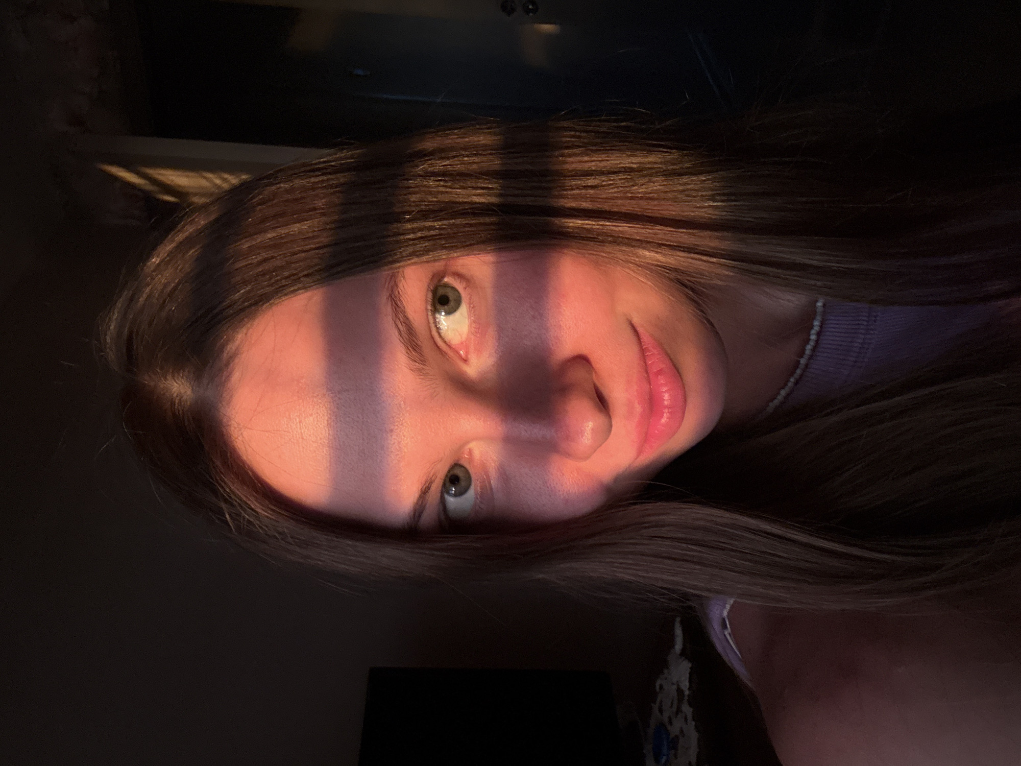 An indoor portrait features a young adult female, illuminated by warm, golden-orange light characteristic of sunrise or sunset. She has fair skin, long straight brown hair parted in the middle, and green eyes, looking directly at the viewer with a slight, closed-lip smile. Distinct horizontal shadows fall across her forehead, eyes, and nose, indicating the light source is partially obstructed, possibly by window blinds or a narrow opening. She wears a light purple ribbed top and a white beaded necklace. The background is largely dark and underexposed, with a dark rectangular screen (likely a television) visible on the left and a doorframe with a door handle faintly discernible on the right. The specific location within the city of Perm, Russia, cannot be determined from the visual information present in the image.