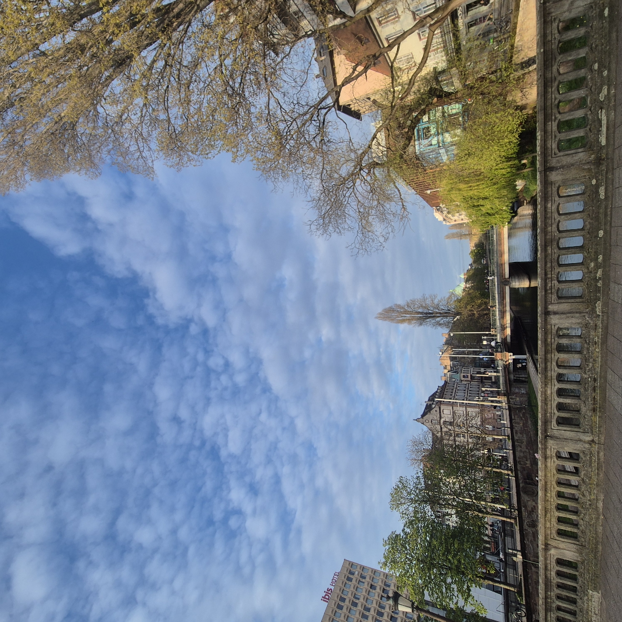 The image shows a view of Strasbourg, France, looking across a canal lined with buildings. The sky is blue with scattered clouds. A stone bridge with decorative openings is visible in the foreground, providing a vantage point over the canal. Buildings, including what appears to be an Ibis hotel, are on one side of the canal, and other buildings with various architectural styles are on the opposite side. Trees with budding leaves line the canal banks. The perspective is slightly angled upward, creating a distorted effect. There are no people prominently visible in the photograph.