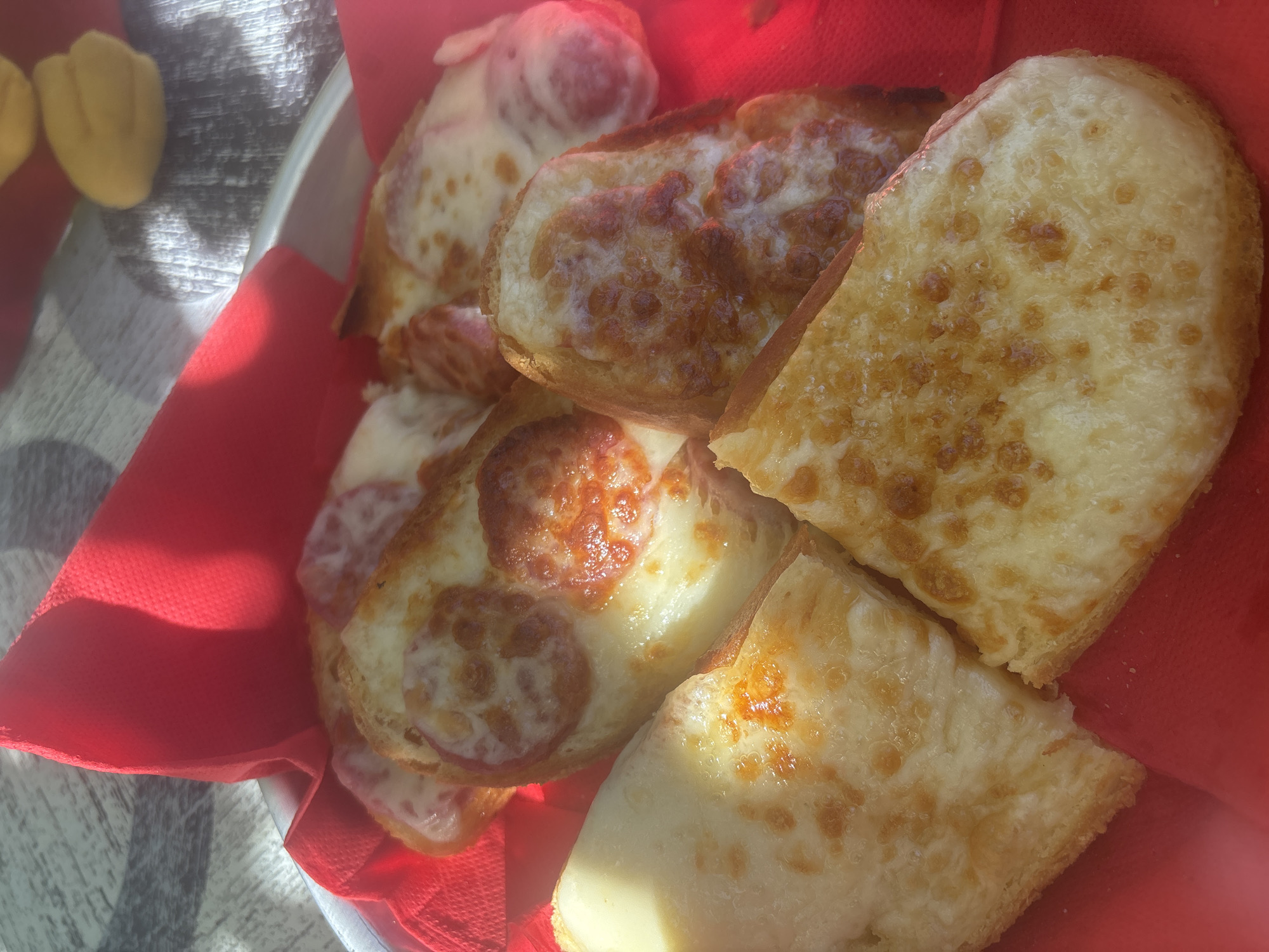 In Trudovets, Bulgaria, a close-up shot captures a serving of open-faced sandwiches, likely bruschetta or mini pizzas, arranged in a silver dish lined with a vibrant red napkin. Several pieces are visible, each topped with melted cheese and some featuring slices of pepperoni. The lighting suggests an outdoor setting, possibly a patio or cafe, with sunlight creating warm highlights and shadows on the food and the textured surface of the table. The overall impression is one of a simple, appealing appetizer.
