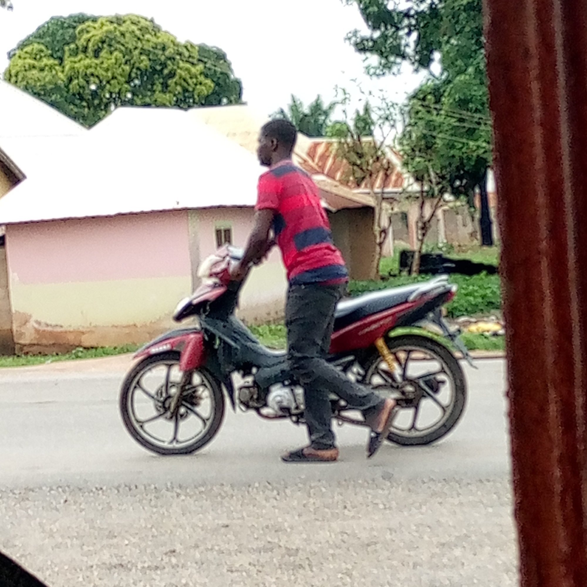 A person is astride a red and black motorcycle on a paved road in Mararaba, Nigeria. The individual wears a red and blue striped shirt, dark trousers, and dark shoes. The buildings in the background have white roofs and colored walls. Lush green trees are prominent around the buildings. The foreground includes a blurry vertical element, potentially part of a structure or vehicle.