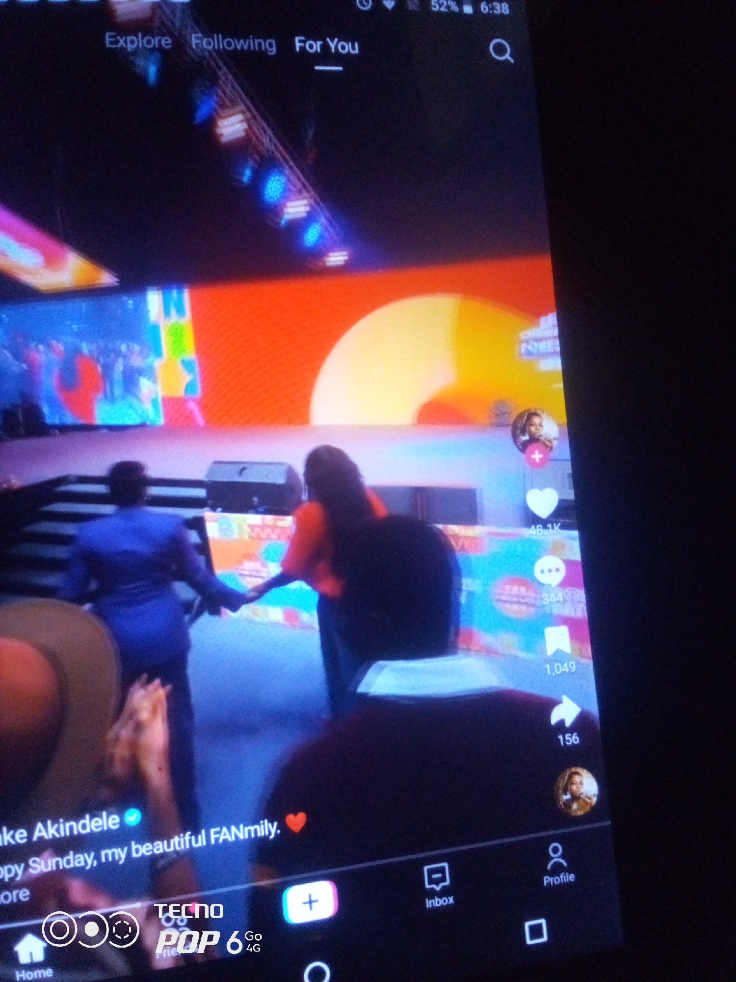 This media file is a screenshot of a mobile device displaying a social media video. The video appears to be taken at an indoor event with a brightly lit stage and a large screen in the background. The screen shows colorful graphics, possibly a logo or animation, with orange and yellow as prominent colors.
In the foreground, two individuals are walking hand-in-hand. The person on the left is wearing a blue suit, and the person on the right is wearing an orange top and dark pants. Their backs are to the camera as they walk away from the viewer, towards the stage. Their postures suggest a shared moment, perhaps a congratulatory walk or a participation in an event. There are other audience members visible in the foreground, some with their hands raised.
On the right side of the screen, there are several interactive icons common to social media platforms, indicating engagement metrics like likes (48.1K), comments (344), shares (1,049), and views (156). A profile picture of a woman is also visible near these icons.
At the bottom of the screen, there is text that reads "ke Akindele Happy Sunday, my beautiful FANmily." This suggests the video is a personal message or update from someone named Akindele, directed towards their fans. The "TECNO POP 6 Go 4G" watermark at the bottom indicates the phone model used to capture or display the screenshot. The time displayed on the phone's status bar is 6:38, and the battery is at 52%. The overall mood seems positive and celebratory, given the bright lighting, the fans' engagement, and the "Happy Sunday" message. The setting appears to be a live event or performance in Nigeria.