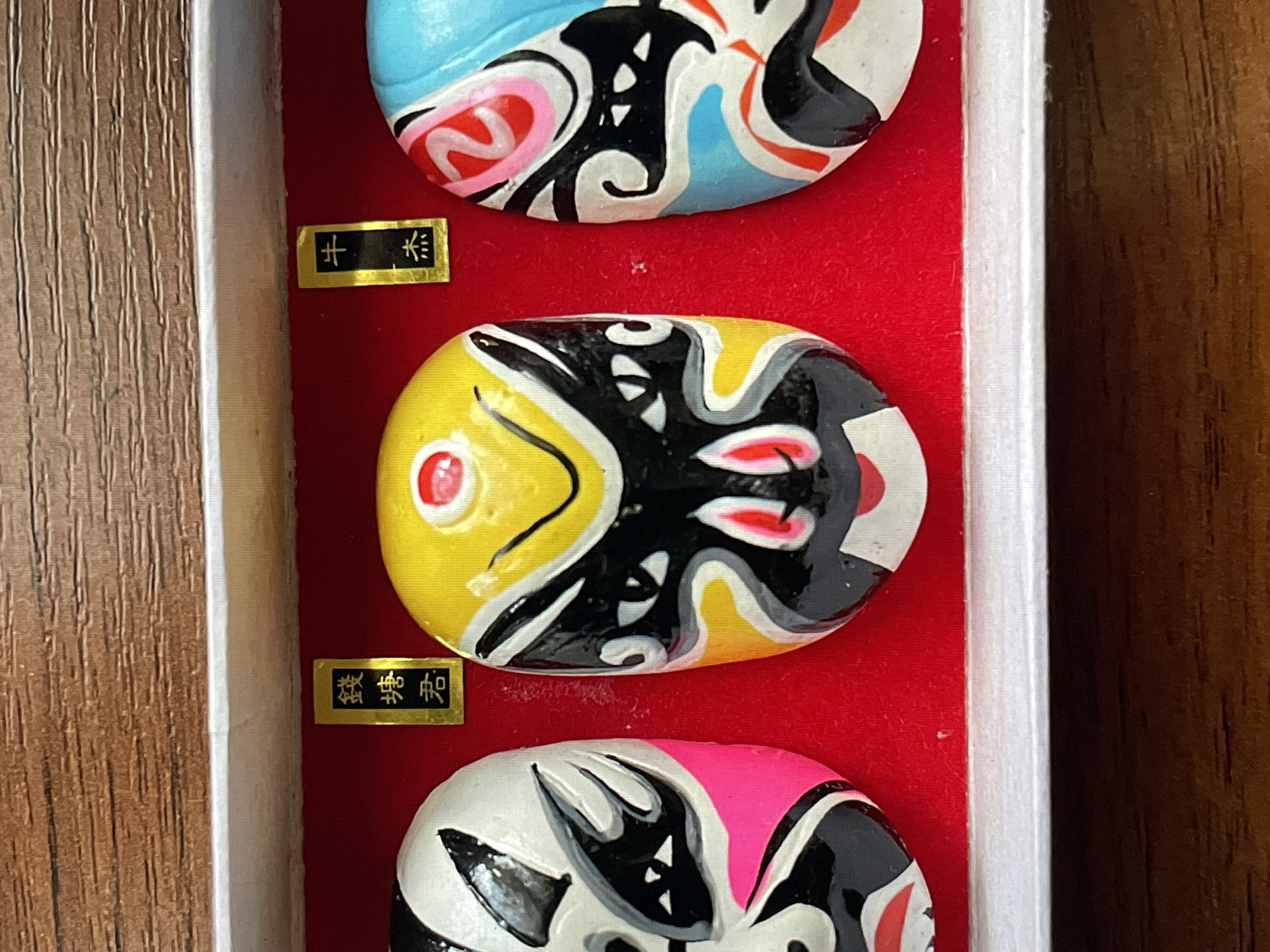 The image displays three miniature, vibrantly colored decorative masks, arranged horizontally within a box. The interior of the box is lined with a textured, deep red fabric, possibly velvet or felt. The upper border of the box is white, and above it, a dark brown surface with a wood grain texture is visible.
From left to right:
1. A partial view of a mask in white, black, and hot pink, featuring stylized facial patterns.
2. A full view of a mask predominantly yellow and black, with white details and distinct red accents, including a circular dot on the forehead and smaller details around the nose and mouth area.
3. A partial view of a mask in light blue, black, and white, with hot pink and red accents, also displaying stylized facial patterns.
Two small, rectangular labels with gold borders and black backgrounds are positioned above the masks. The label above the left mask shows vertical white Chinese characters that read "鐵塘君" (Tiě Táng Jūn). The label above the right mask shows vertical white Chinese characters that read "牛杰" (Niú Jié). No people are visible. The environment appears to be an indoor setting. The location in the city of Saint Petersburg, Russia, cannot be determined from the image provided, as the depicted objects are culturally Chinese in origin.