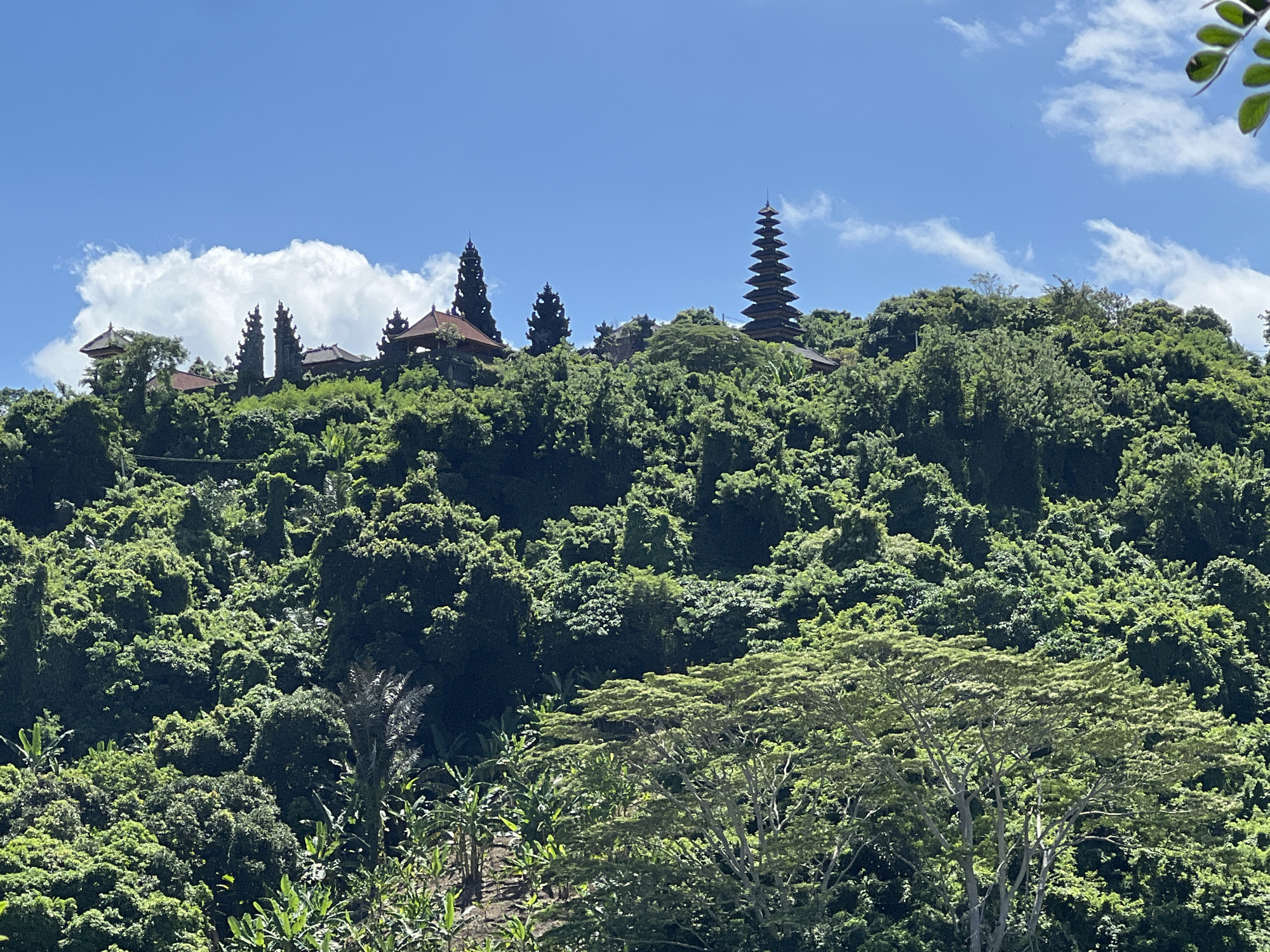 The image depicts a hillside densely covered with lush, green vegetation. At the top of the hill, several buildings with traditional Balinese architecture are visible, including tiered pagodas. The sky is bright blue with scattered white clouds. The location appears to be Dawan, Indonesia, based on the architectural style and tropical environment.