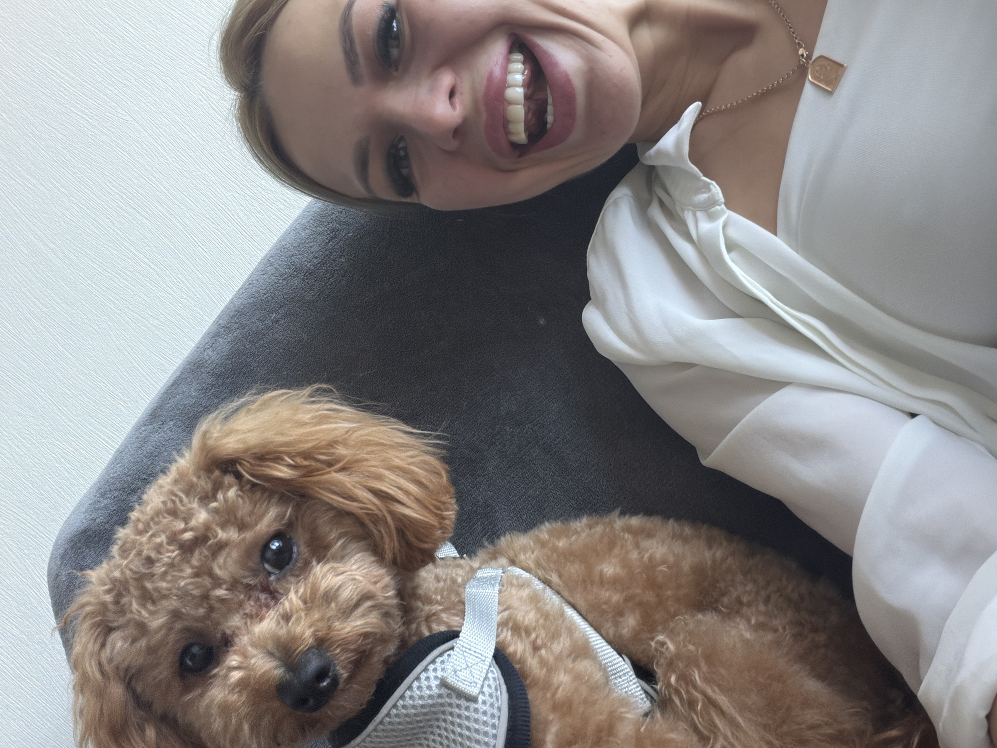 In a close-up selfie, a woman with blonde hair, wearing a white blouse and a necklace with a pendant, is smiling broadly, her mouth open. Beside her, a small, tan poodle wearing a grey harness is looking at the camera. They are both seated on a dark grey sofa. The background is a textured light-colored wall. The setting appears to be indoors, likely in a residential environment within the city of Kyiv, Ukraine.
