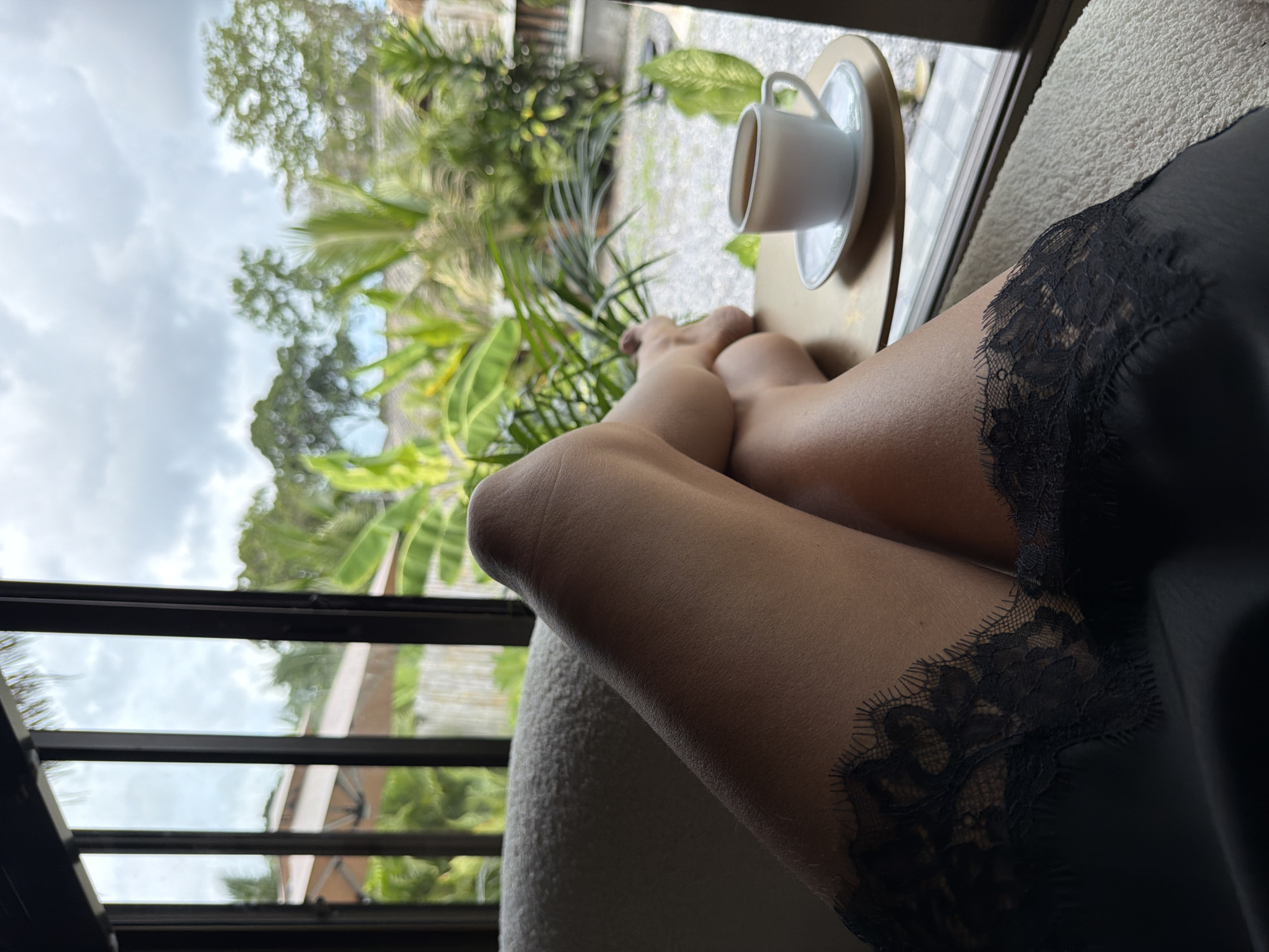 An interior shot shows a person's legs crossed, resting on a textured cream-colored sofa, with a view of a tropical outdoor scene through a large window. The person is wearing black lace-trimmed attire. On a small, round, brass-colored table beside the sofa, a white cup of coffee sits on a saucer. The background outside the window reveals lush greenery, palm trees, and a cloudy sky, suggesting a humid, tropical climate. The architectural elements visible through the window include what appears to be a thatched roof and wooden structures, indicative of a resort or villa in Baan Bo Phut, Thailand. The lighting suggests an overcast day, with soft natural light filling the room.