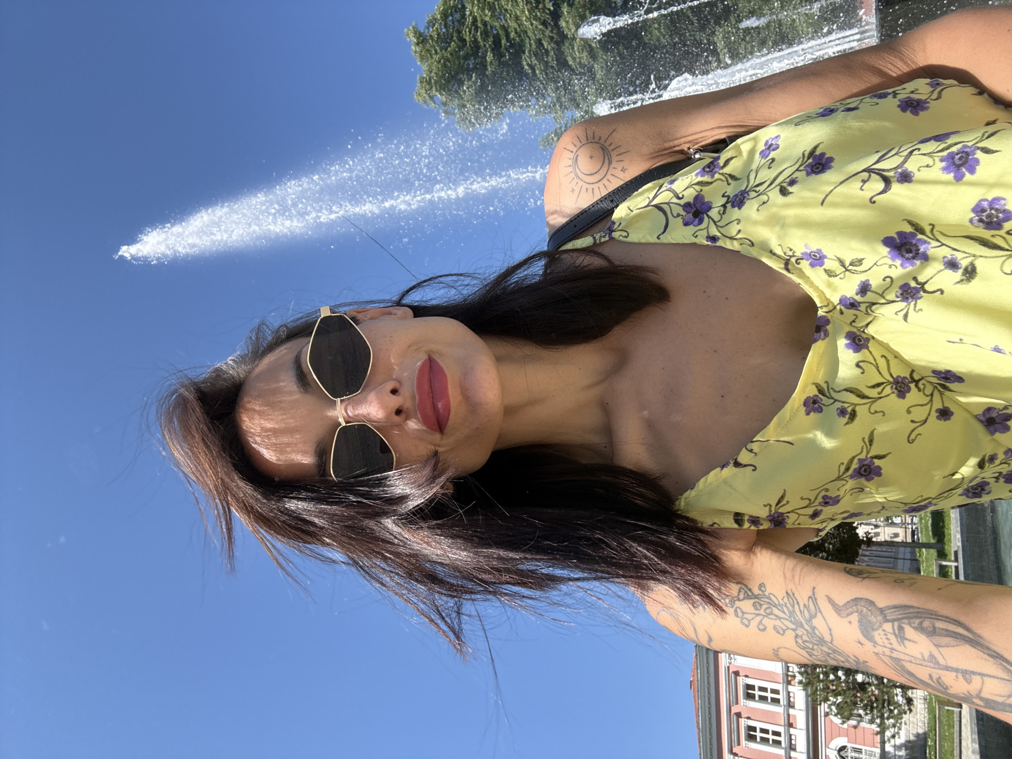 A woman wearing sunglasses and a yellow floral dress is taking a selfie in front of the water fountain in the City Garden in Pleven, Bulgaria. The background features a blue sky, the fountain's water spray, trees, and a building with white-framed windows. The woman has dark hair and visible tattoos on her arm and shoulder.