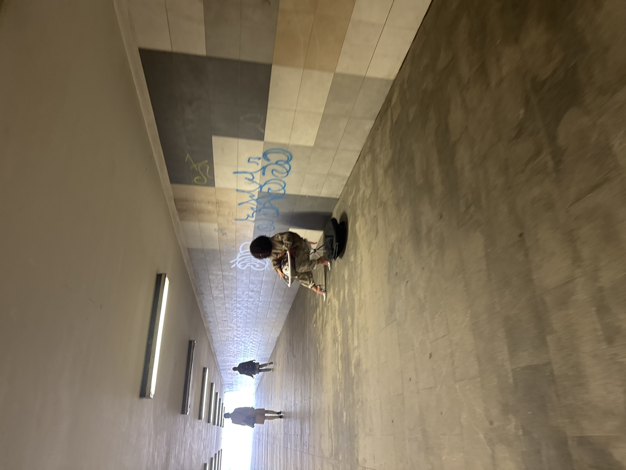 The image depicts the interior of a pedestrian tunnel in Riga, Latvia. The tunnel has walls tiled with a pattern of light and dark grey rectangles, some sections of which are covered in graffiti. A musician is positioned to the right of the tunnel, playing a steelpan instrument while seated on a stool. A musical instrument case is on the floor near the musician. Two pedestrians are walking further down the tunnel, away from the camera. Recessed strip lights line the left side of the tunnel. The overall environment is dimly lit with a bright light coming from the end of the tunnel, giving the scene an artificial glow.