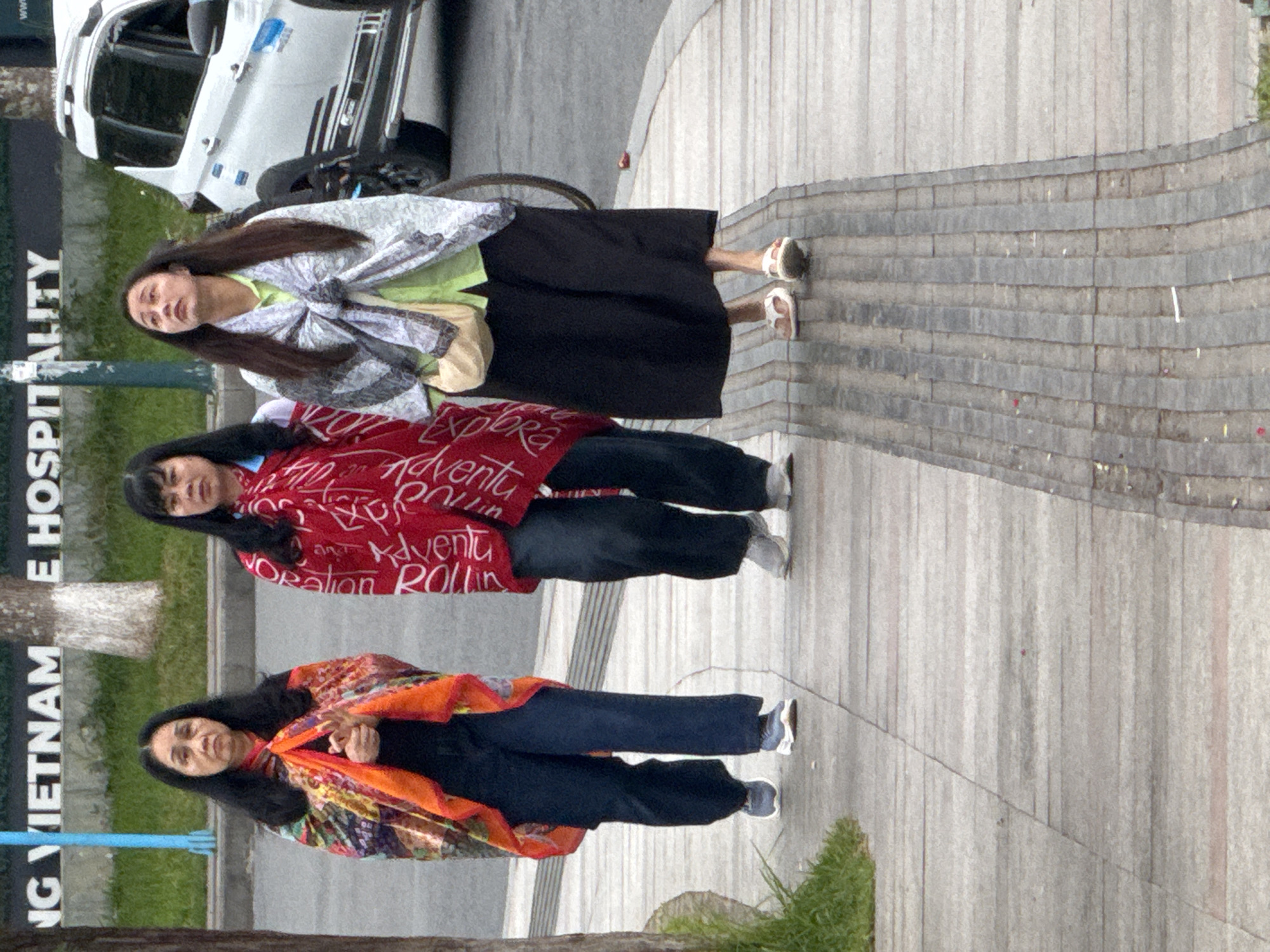 Three Vietnamese women are walking on a sidewalk in front of a street. The woman on the left is wearing a colorful patterned scarf over a black top and dark blue pants, and sneakers. The woman in the middle is wearing a red blanket-like garment with white writing over a dark top and pants, and sneakers. The woman on the right is wearing a light green shirt and dark pants, a patterned scarf, and white sandals. She is looking up. Behind them, a white vehicle and a bicycle wheel are visible. The background shows a building with the words "NG VIETNAM E HOSPITA" visible on it. The sidewalk is made of concrete slabs with tactile paving. This scene is consistent with an urban environment in Da Lat, Vietnam, possibly near a hospital or a large institution.