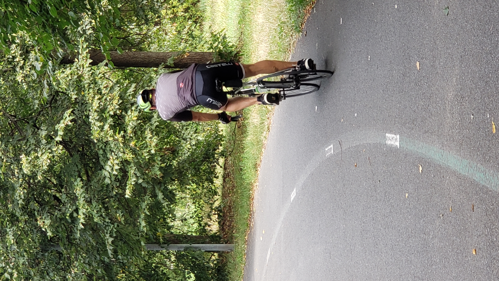 A person is cycling on a paved path lined with trees and grass. The cyclist, wearing a helmet, cycling jersey, and shorts, is viewed from the rear as they pedal away from the camera. The path has white dashed lines painted on it, indicating a lane division. The environment appears to be a park or a recreational area in Prešov, Slovakia.
