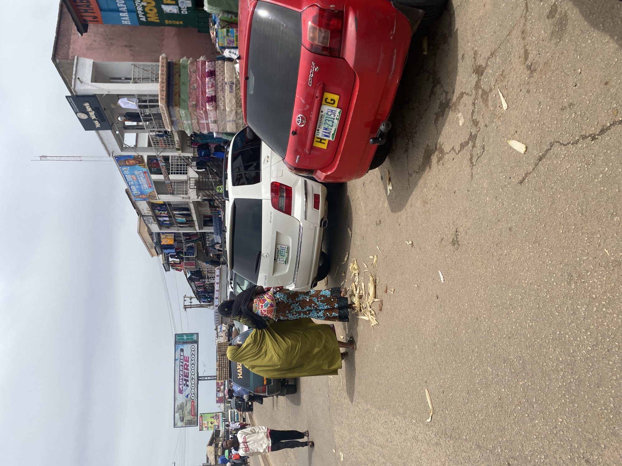 In Jos, Nigeria, a street scene unfolds with a bustling marketplace atmosphere. The foreground features a red Toyota Corolla with a yellow license plate reading "WAN-2315H G". To its left, a white Mercedes-Benz SUV, identified as a GLK 350 4MATIC, is parked. Two women are walking on the asphalt road; one, in a flowing green hijab, is accompanied by another woman wearing a patterned skirt and a black headscarf, with a child wrapped around her back. Scattered on the ground are remnants of what appear to be corn husks.
In the background, a row of shops lines the street, displaying various goods. A large billboard, promoting advertising services with the text "ADVERTISE HERE" and a phone number, is prominently visible. Further down, shops display clothing and other merchandise. Traffic, including cars and possibly motorcycles, is present, suggesting a busy urban environment. The sky is overcast, indicating a cloudy day. The overall scene captures the vibrant commercial activity characteristic of a Nigerian city street.