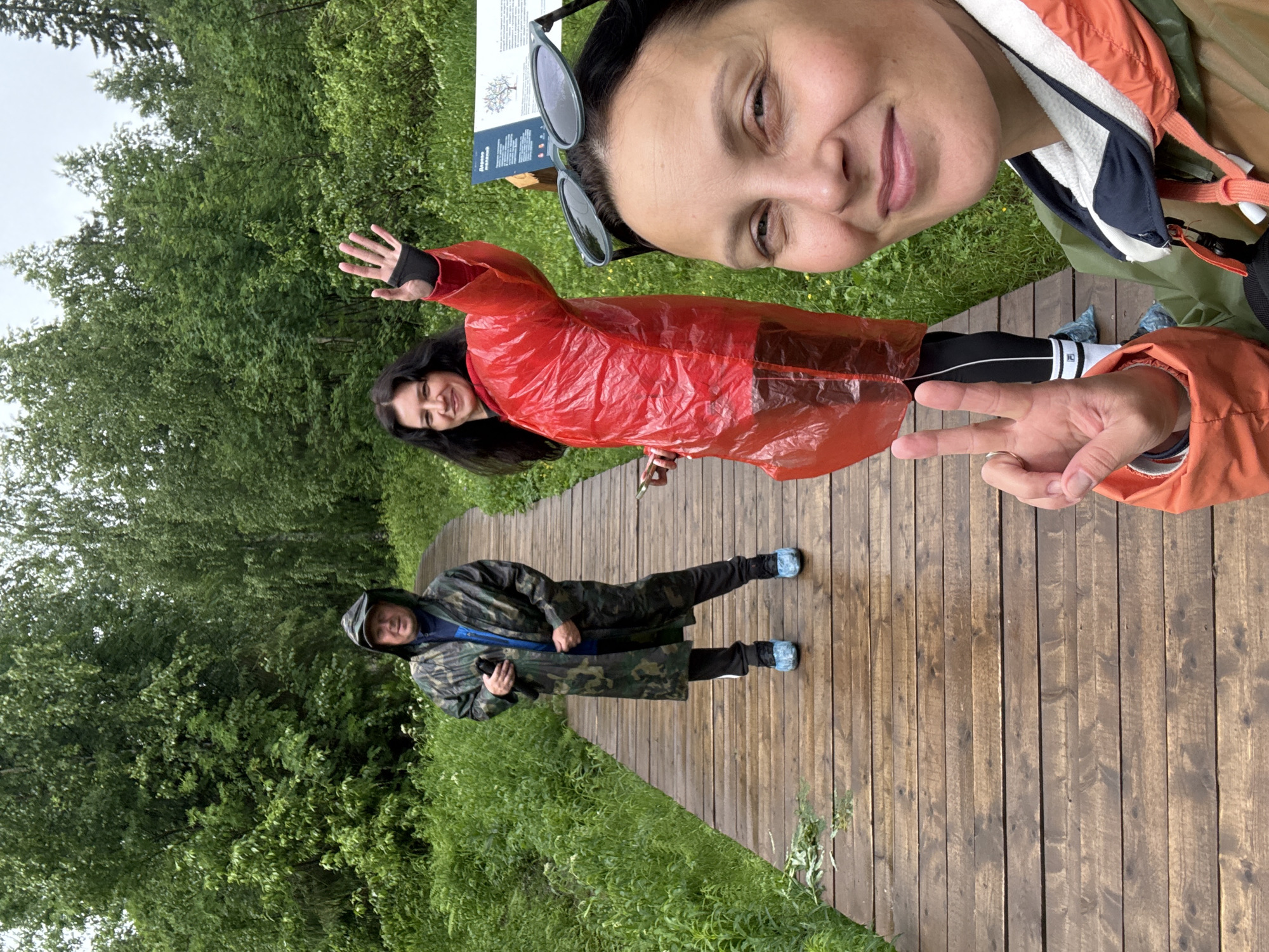 Three people are walking on a wooden boardwalk in a forest. The person in the foreground is taking a selfie and making a peace sign with their fingers. The person in the middle is wearing a red raincoat and waving. The person in the background is wearing a camouflage raincoat. The boardwalk appears to be wet, suggesting it may have recently rained. The surrounding environment is lush with green trees and foliage. The location is the boardwalk to the Usvinskaya Stone geological monument in the city of Usva, Russia.