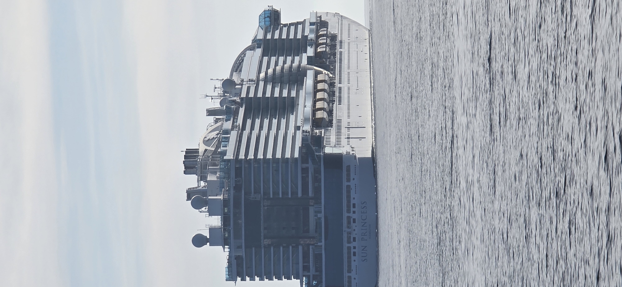 The image displays a large cruise ship, identified as the "SUN PRINCESS", navigating open waters under an overcast or hazy sky. The vessel is oriented horizontally within the frame, showcasing its immense scale and modern design. Its superstructure is a mix of white and various shades of grey, featuring multiple decks lined with numerous private balconies. Along the sides of the ship, several rectangular lifeboats are visible, ready for deployment. The top decks reveal intricate architectural elements, including curved glass enclosures and a cluster of communication antennas and radar domes.
The name "SUN PRINCESS" is clearly legible in large letters on the ship's dark blue hull, with smaller, lighter text underneath that appears to read "MAJURO," indicating its port of registry. The water surrounding the ship is a uniform grey-blue, exhibiting a subtly rippled or choppy texture, suggesting either movement or a gentle breeze. The sky above is a pale, uniform light blue-grey, indicative of daytime with significant cloud cover or haze. There are no discernible human figures or specific activities taking place, creating a serene and expansive maritime scene. Given the context, the ship is located in or near Bar, Montenegro.