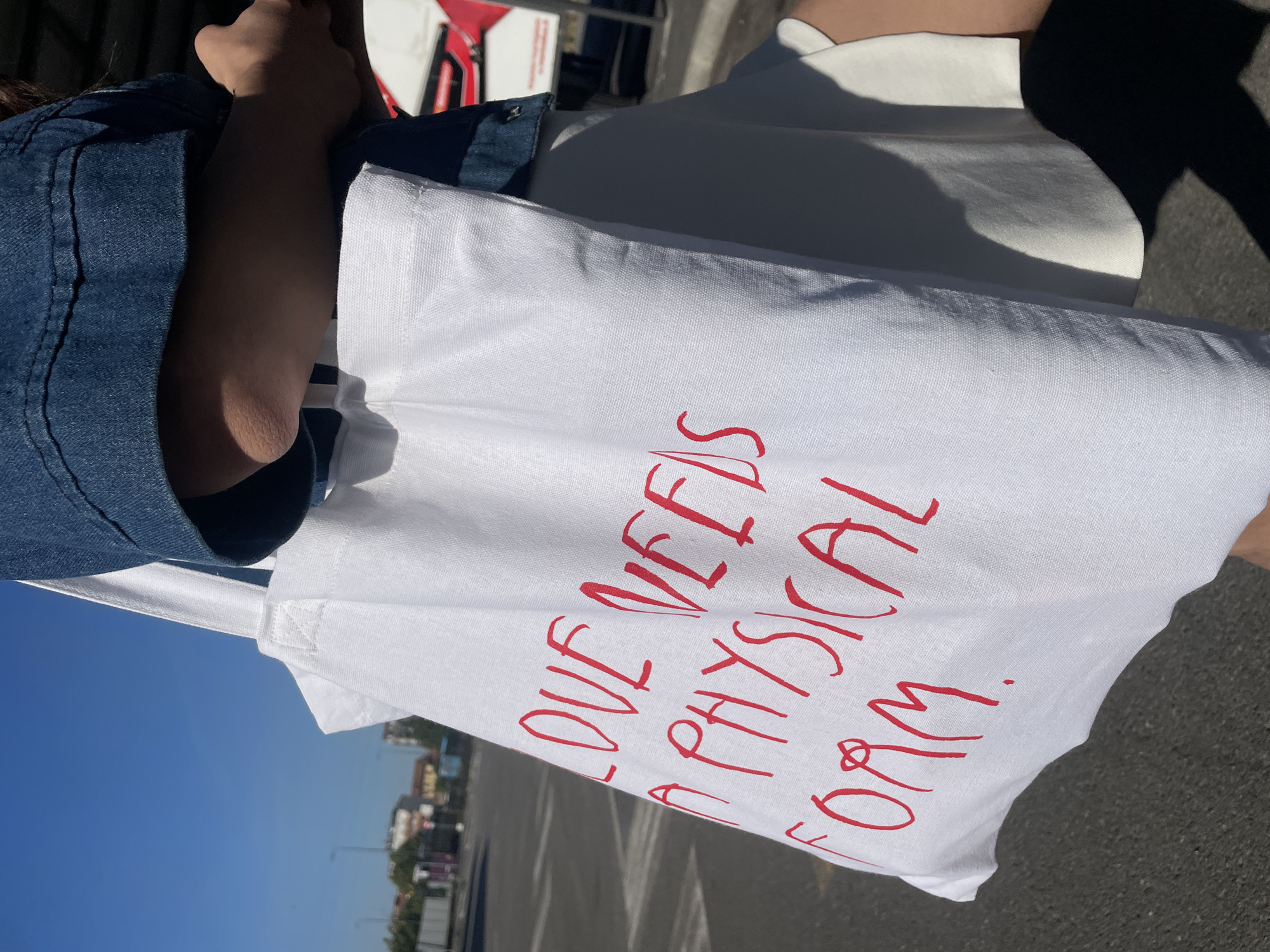 A person wearing a denim jacket and white skirt is holding a white tote bag with the words "LOVE NEEDS A PHYSICAL FORM." written in red. The photo appears to be taken outdoors on a clear day. The location is likely Milan, Italy, given the architectural style of buildings in the background.