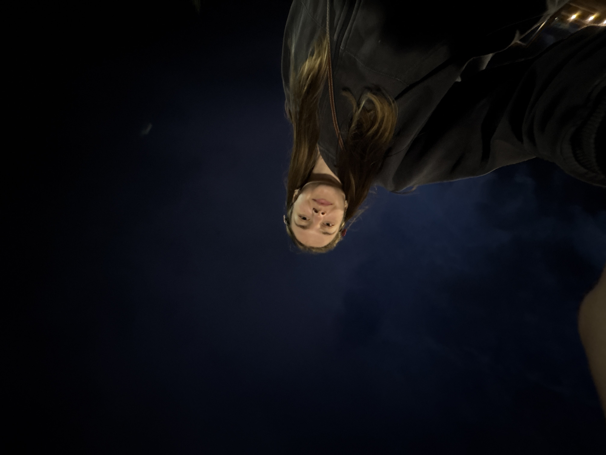 The image, captured at night, shows a young woman with light brown hair looking up at the camera. She is wearing a dark-colored hooded sweatshirt and a strap from a bag or purse is visible over her shoulder. The background is a dark, night sky, with a faint crescent moon visible in the upper right quadrant. There is a hint of smoke or mist in the lower left portion of the image. The perspective suggests the photo was taken from a low angle. The location is stated to be in the city of Nizhniye Mulli, Russia.
