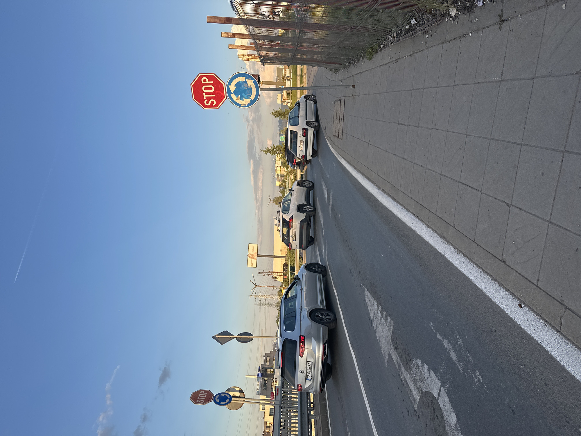 The image depicts a street scene in Sofia, Bulgaria. The foreground shows a grey asphalt road with faded white directional arrows, bordered by a white painted line. The sidewalk to the right is paved with square grey tiles. A metal fence runs alongside the sidewalk.
A row of parked cars lines the road, including a silver Volkswagen Passat with the license plate "CB 4136 XA." Stop signs and roundabout signs are visible, along with other traffic signage, indicating a possible intersection or roundabout.
The background includes buildings and industrial structures, a billboard, and power lines. The sky is clear blue with hints of clouds.