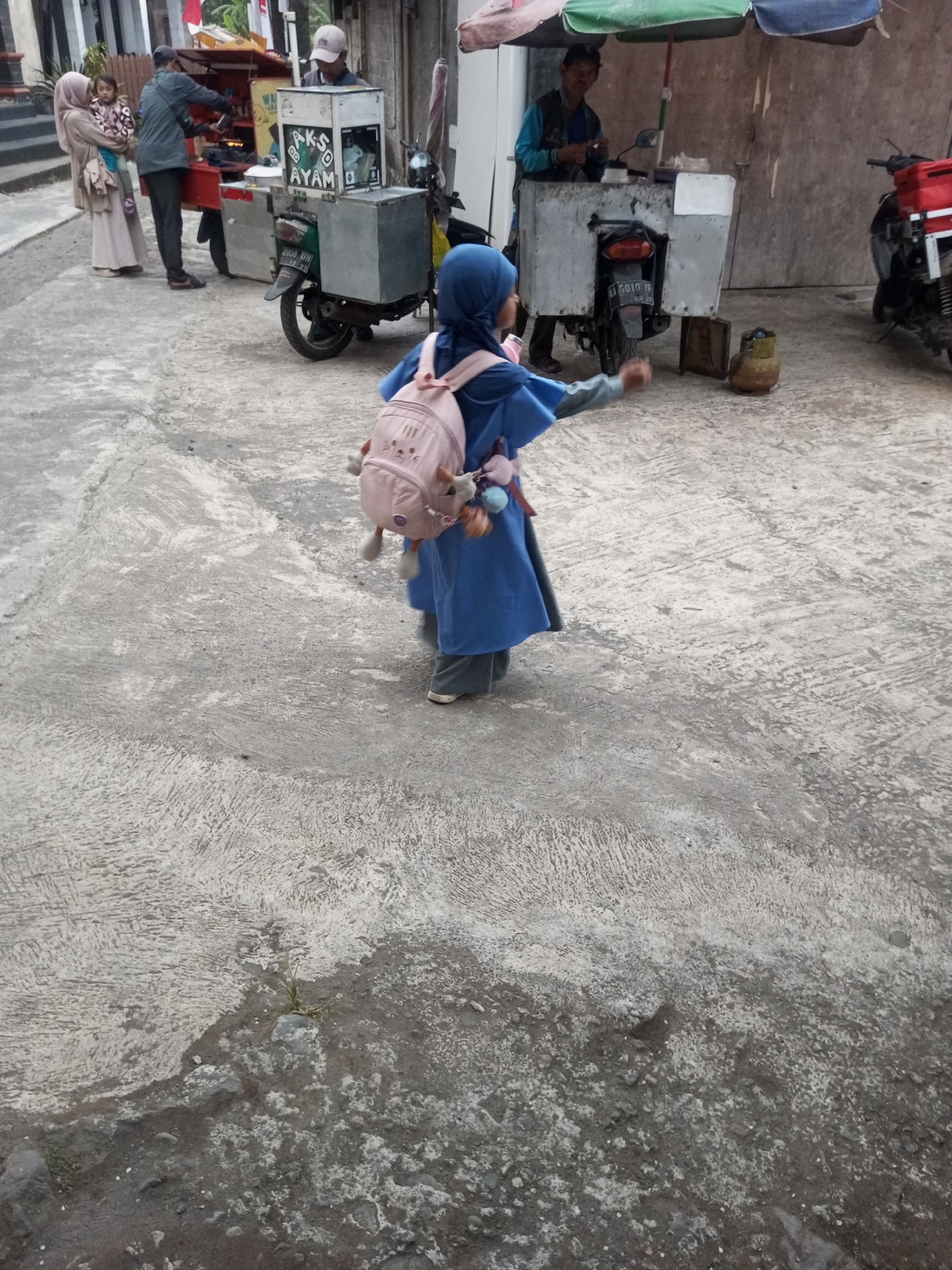 In a street scene in Kalibeber, Indonesia, a young girl wearing a blue hijab and a matching blue dress, carrying a pink backpack adorned with stuffed animal keychains, stands facing away from the camera and gestures with her right hand. She is positioned in the foreground on a concrete ground with visible water stains and textures. Behind her, a food cart with a motorcycle chassis is parked. Two individuals are visible at the food cart: a man wearing a blue shirt and a cap is operating the cart, and another man in a blue shirt and a cap stands to the right of the cart. To the left of the food cart, another person is holding a child. A red motorcycle with a storage box is parked to the right of the scene. The overall environment appears to be an outdoor commercial area with food vendors.