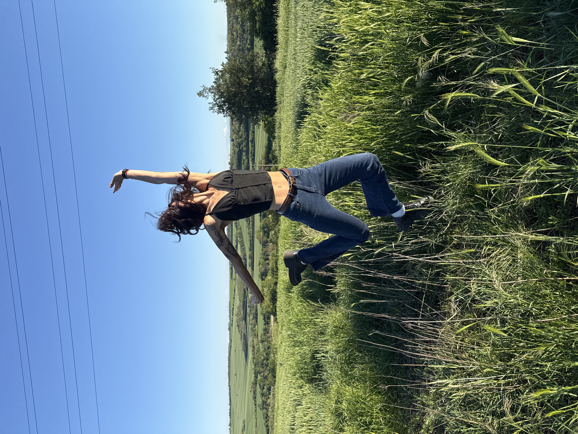 The image presents a young adult female in an outdoor field setting. She is wearing a black top, blue jeans, and brown footwear. Her posture suggests a dynamic movement, perhaps a dance or jump, with one arm raised towards the blue sky and the other extended. The environment includes tall green grass, a visible landscape in the background with trees and fields, and a clear blue sky. Power lines are visible in the upper left corner of the frame. There's no information to suggest this image was taken in Pleven, Bulgaria. The image does not contain any buildings or landmarks associated with the city.