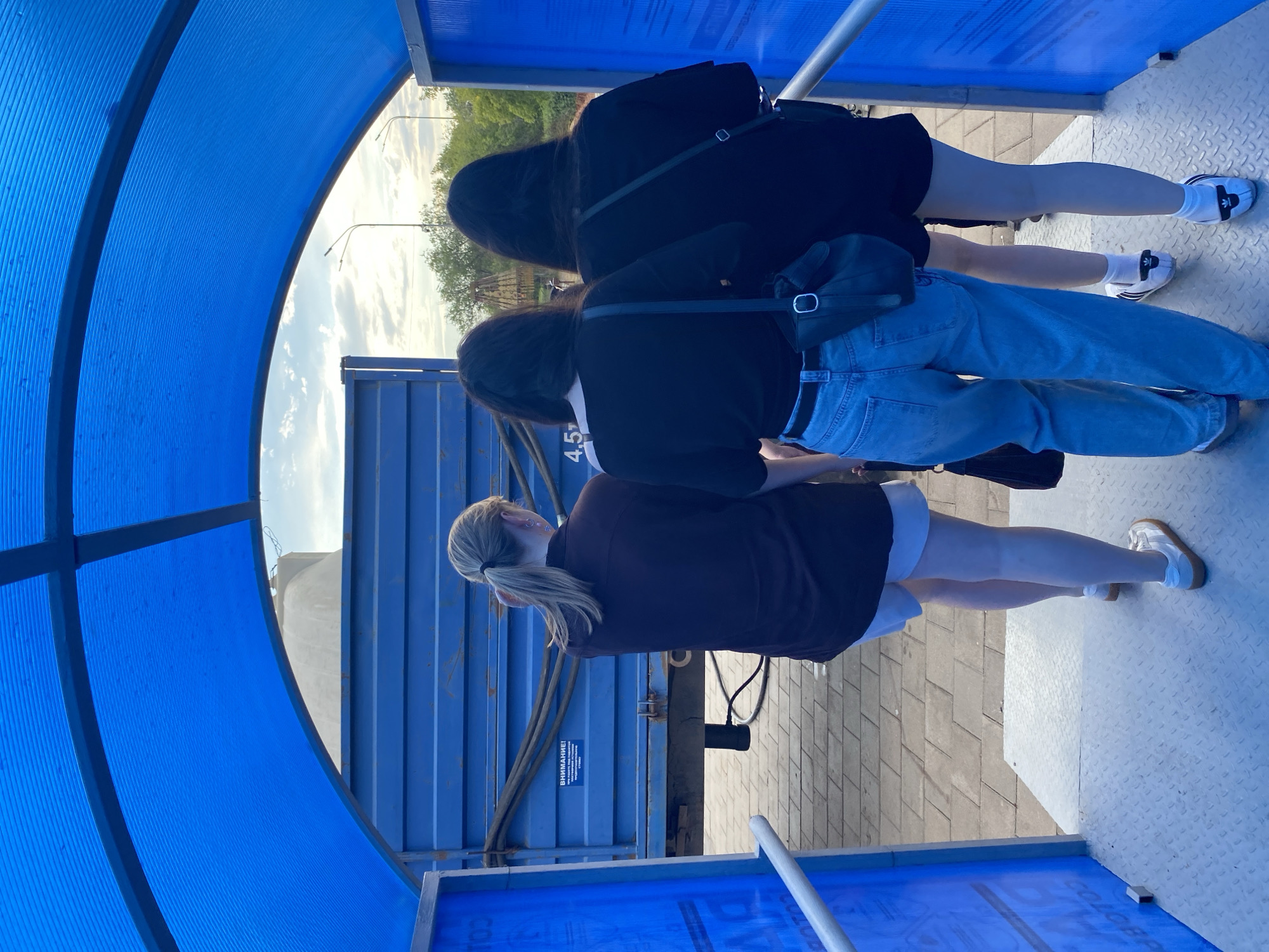 Three individuals, viewed from behind, stand beneath a blue arched canopy. The person on the left has blonde hair in a ponytail and wears a dark maroon shirt and white shorts. The middle person has dark hair and wears a black t-shirt and light blue jeans, carrying a black crossbody bag. The person on the right also has dark hair, wears a black t-shirt and black shorts, and carries a black crossbody bag. In the background, a blue truck with the text "4.5t" and some warning signs is visible. The ground is paved with grey interlocking tiles and a metal walkway. The sky is cloudy with sunlight. The scene appears to be an outdoor loading or unloading area.