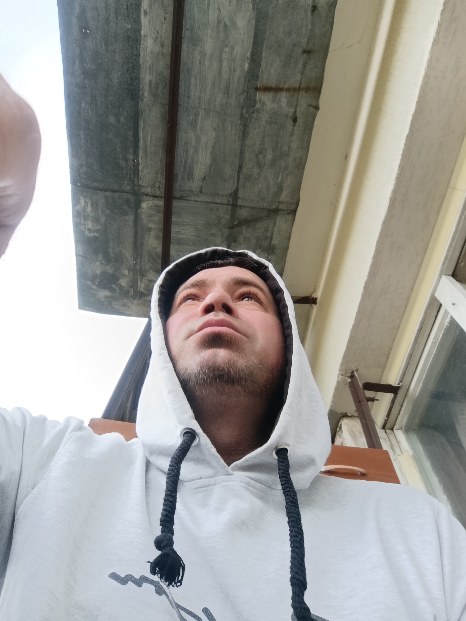 A low-angle, wide shot captures a Caucasian male, appearing to be in his late 30s or early 40s, looking upwards and to the right, with his face partially obscured by a white hooded sweatshirt. The man has a fair complexion, short brown hair, and is clean-shaven apart from a light stubble on his chin and jawline. He is wearing the hood of his sweatshirt up, and a black braided drawstring with a tassel hangs down the front of the garment. The sweatshirt has a gray graphic design on the lower chest area.
The background suggests an outdoor setting, possibly a porch or balcony. Above and behind the man's head is a gray, weathered overhang, likely made of concrete or metal, supported by a dark brown metal pole. To the right of the man, a cream-colored building facade is visible, with textured plasterwork and a white-trimmed window with visible panes. The overall lighting is overcast, with a bright sky peeking through the overhang on the left. The image appears to have been taken in Ladyzhyn, Ukraine, based on its context.