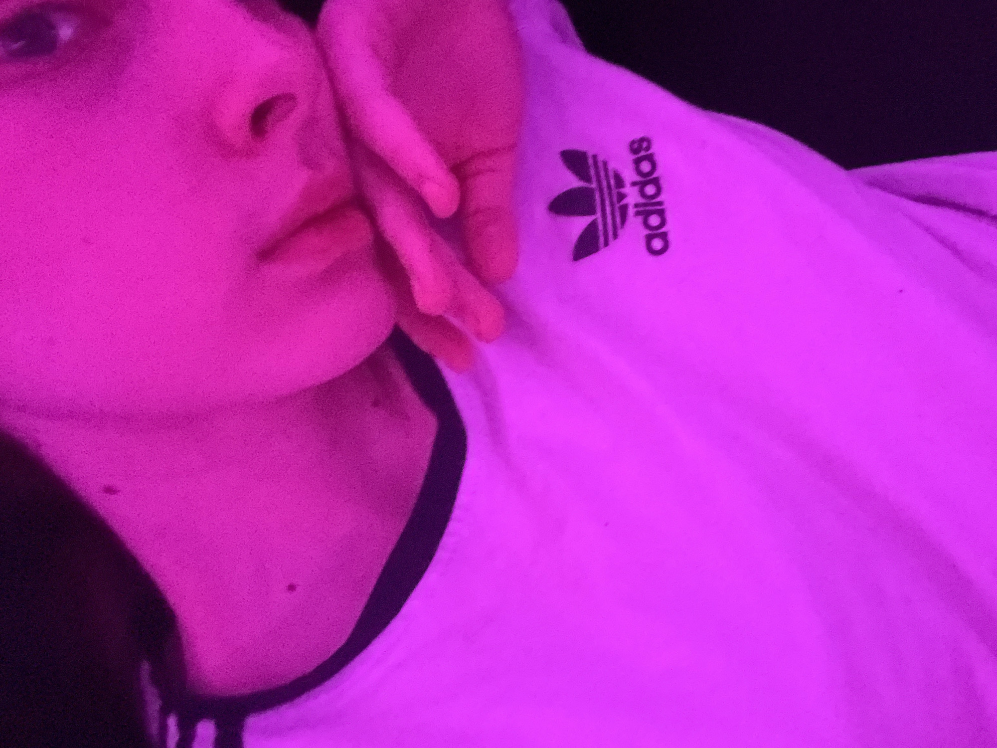 The image captures a close-up of a person bathed in strong fuchsia or magenta lighting, creating a vibrant and somewhat mysterious atmosphere. The subject's face is partially visible, showing one eye, the bridge of the nose, and lips, all heavily tinted by the pink light. A hand with long fingers rests gently against the side of their face or neck, also illuminated in the same intense color.
The person is wearing a white t-shirt or top with black trim along the collar or neckline. Prominently displayed on the chest area of the shirt is a black Adidas Trefoil logo, positioned vertically, with the brand name "adidas" written alongside it. The background is dark and out of focus, suggesting a low-light indoor setting. The intense, artificial lighting obscures natural skin tones and gives everything a surreal, dreamlike quality. The overall mood is calm and contemplative, possibly a self-portrait or a posed shot in an ambient setting. The image has a slightly grainy texture, which adds to its atmospheric quality.