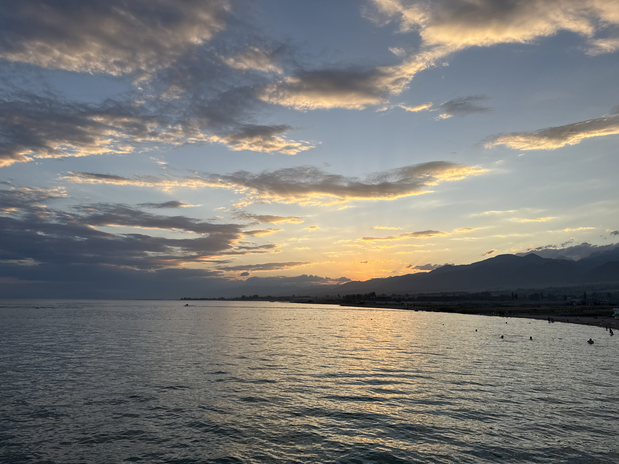 This is a wide-angle, horizontal photograph of a serene lake scene in Sary-Oy, Kyrgyzstan, likely captured during sunset or sunrise. The sky is a dominant feature, painted with a gradient of blues and oranges, with scattered clouds catching the warm light. The sun, low on the horizon, casts a golden glow across the water, creating shimmering reflections.
The foreground is filled with the calm, rippling surface of the lake. In the middle ground, a narrow strip of land is visible, lined with trees and some buildings, suggesting a populated area along the shore. Several people are scattered along this shoreline, and a few are in the water, appearing to swim or wade. The distant background is dominated by the silhouette of dark, majestic mountains, adding a dramatic backdrop to the tranquil scene. A small boat can be seen on the water in the distance. The overall impression is one of peaceful natural beauty and a leisurely atmosphere.