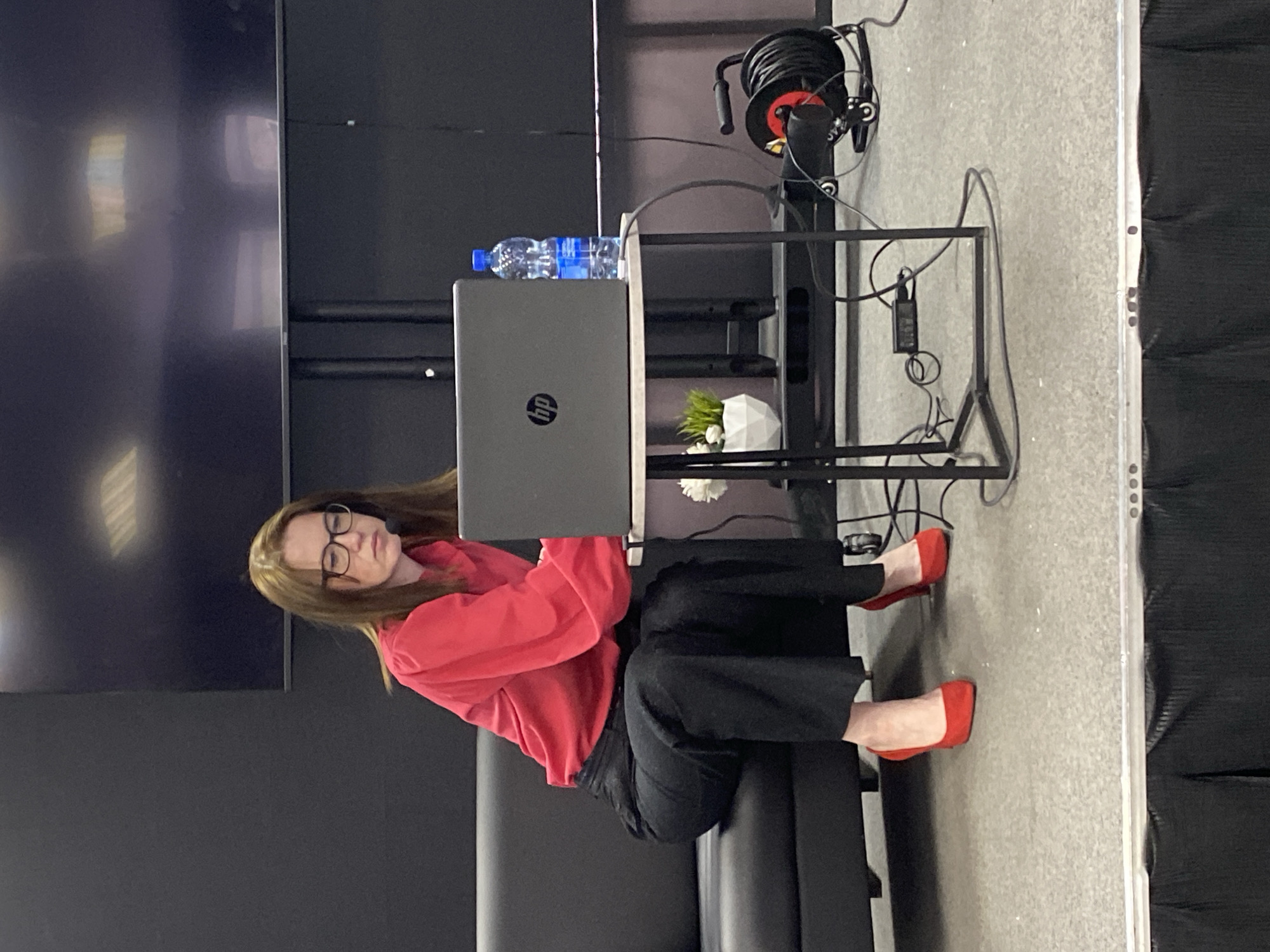 A woman with light brown hair and glasses is seated on a black sofa, positioned in front of a black metal table that holds an HP laptop, two bottles of water, and a small, white potted plant. She is wearing a red sweater and black pants, with red shoes. The backdrop consists of a large black screen on the left and a light gray wall on the right. A cable reel is visible on the right side of the table. The location is unidentified.