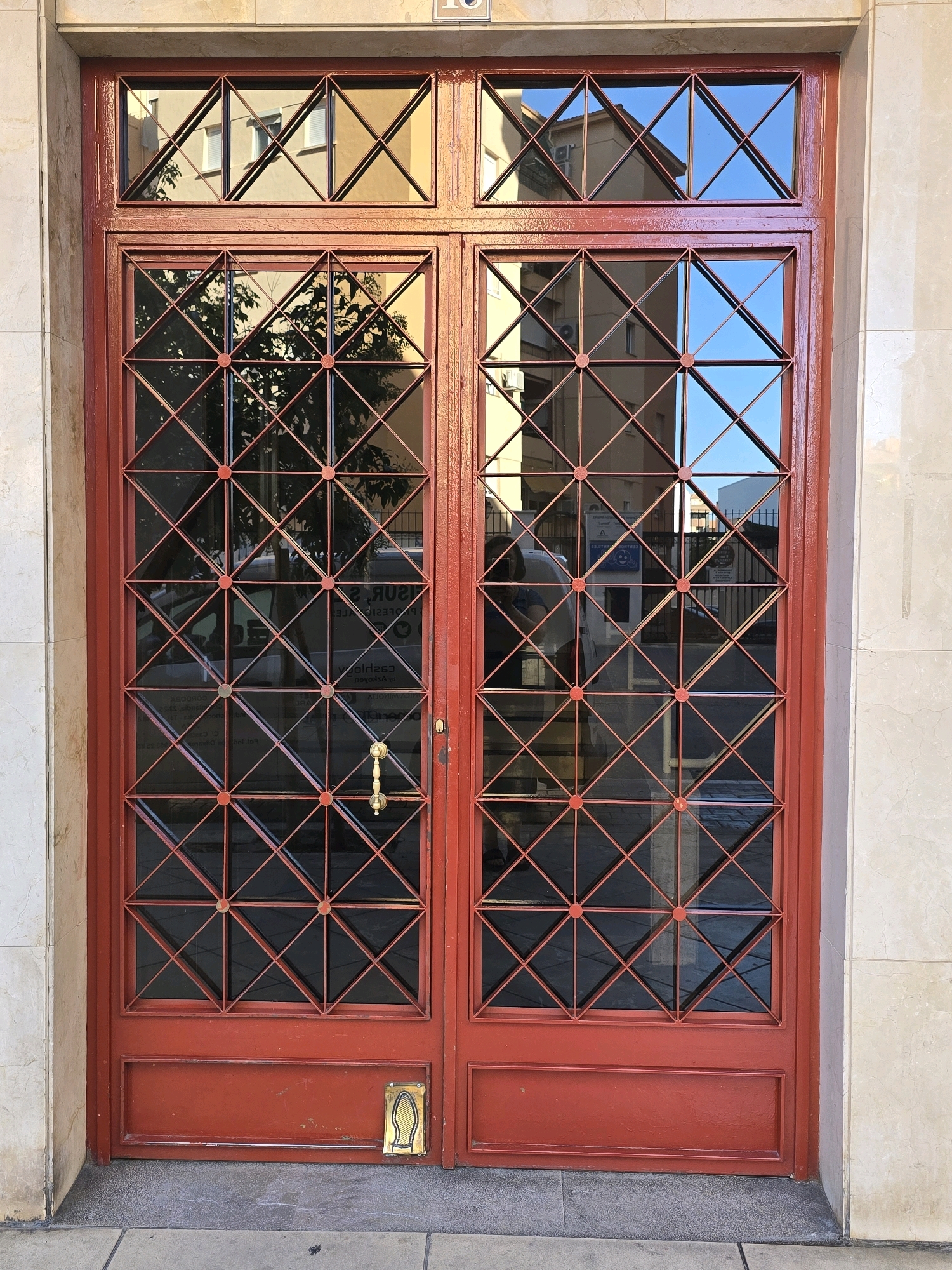 The image shows a red, double-door entrance with a geometric metal grate over the glass panels. The grate is made of interconnected squares, each divided by diagonal lines into four triangles, with small circular connectors at the intersection points. The doors are set within a light-colored stone building facade. The glass panels reflect the surrounding environment, including buildings across the street, a tree, and the sky. A gold-colored door handle is visible on the left door. At the bottom of the right door, there is a small, gold-colored plate with an embossed image of a footprint. The scene appears to be on a street in Jaén, Spain, as suggested by the architectural style and lighting.
