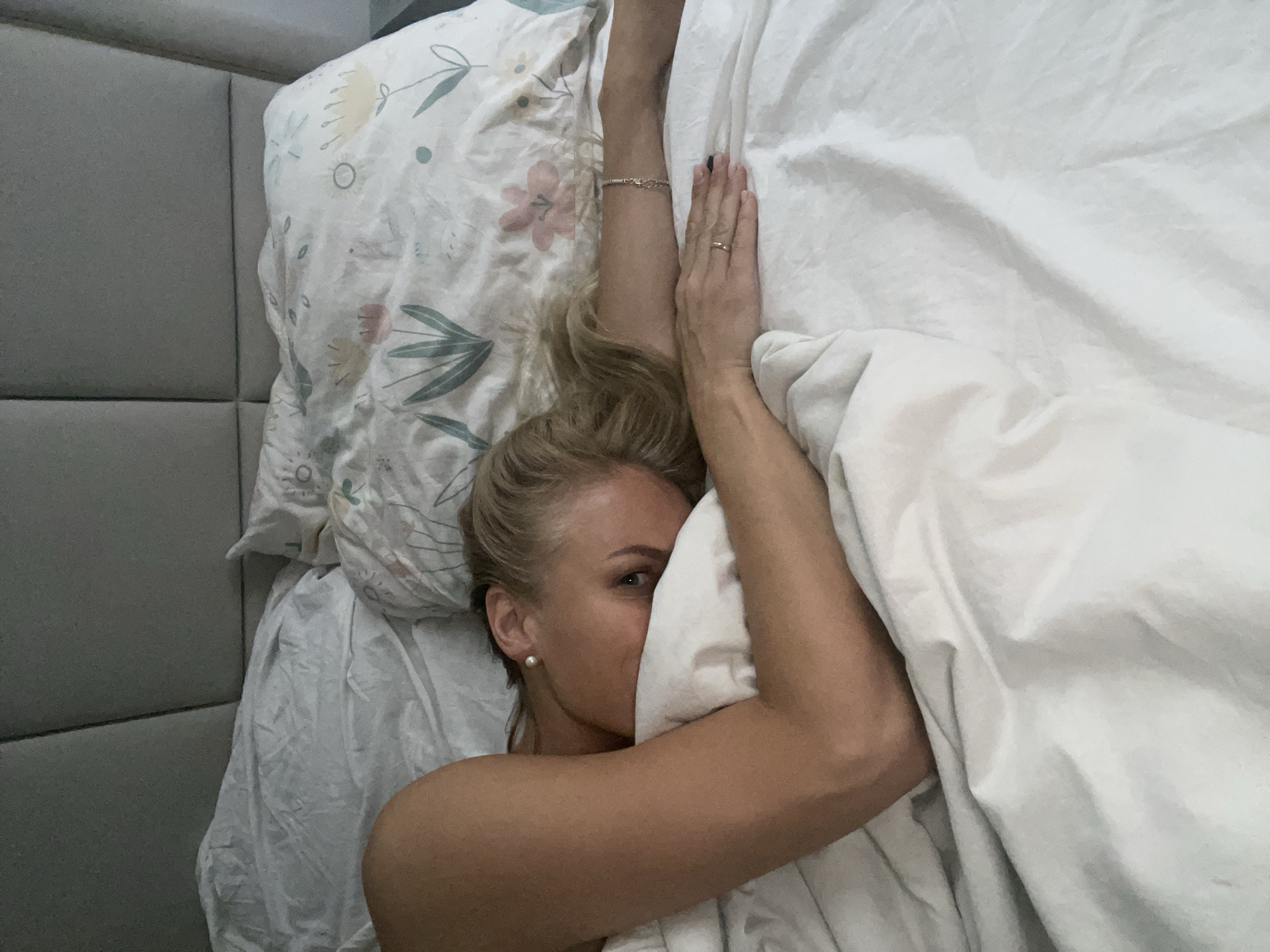 A woman is lying in bed, partially covered by a white sheet, with her face partially obscured by the sheet. She is wearing a pearl earring and a bracelet on her arm. Her blonde hair is swept back from her face. The bed has a floral-patterned pillow and a gray headboard. The location in the city of Baranovka, Russia is not evident in the image.