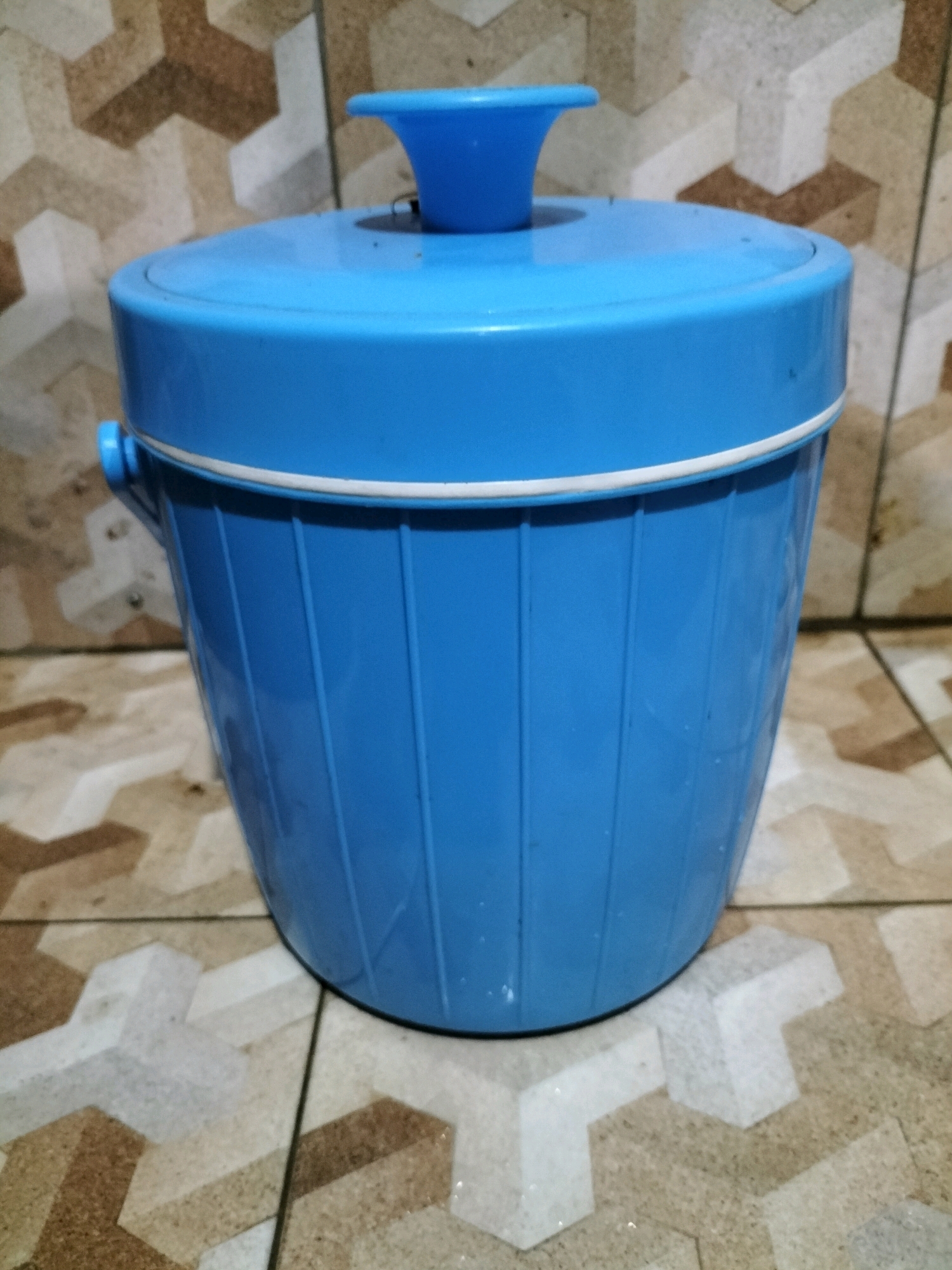 A blue plastic ice bucket with a white rim and a blue lid with a knob is positioned on a tiled floor. The tiles have a geometric pattern in shades of brown, beige, and white. The background appears to be a wall with similar geometric tiling. The scene is likely indoors, possibly in a domestic setting or a small shop, in Tasikmadu, Indonesia. There are no people visible in the image.