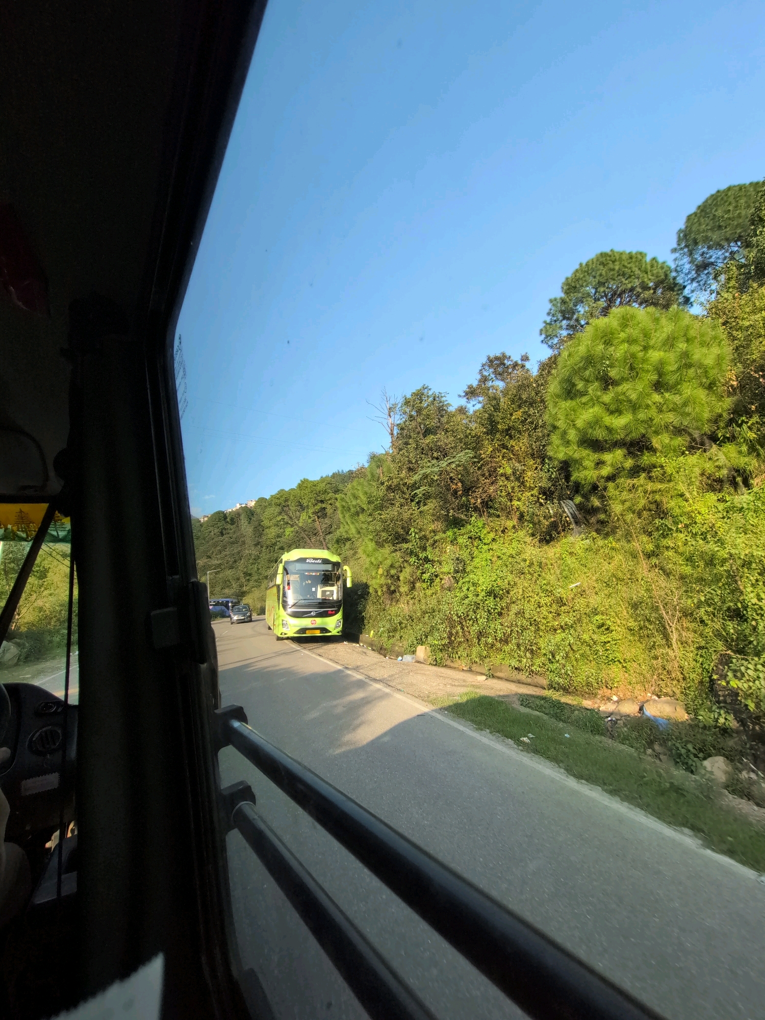 The image is taken from inside a vehicle, looking out through the window at a road scene in Dharamshala, India. The sky is clear blue. On the road, a bright green bus is traveling away from the viewer, with other vehicles behind it. The road is bordered by lush green vegetation and trees, with a hillside visible in the background. Some of the vegetation appears to be pine trees. The sunlight suggests it is daytime. The overall impression is of a journey through a scenic, hilly region.