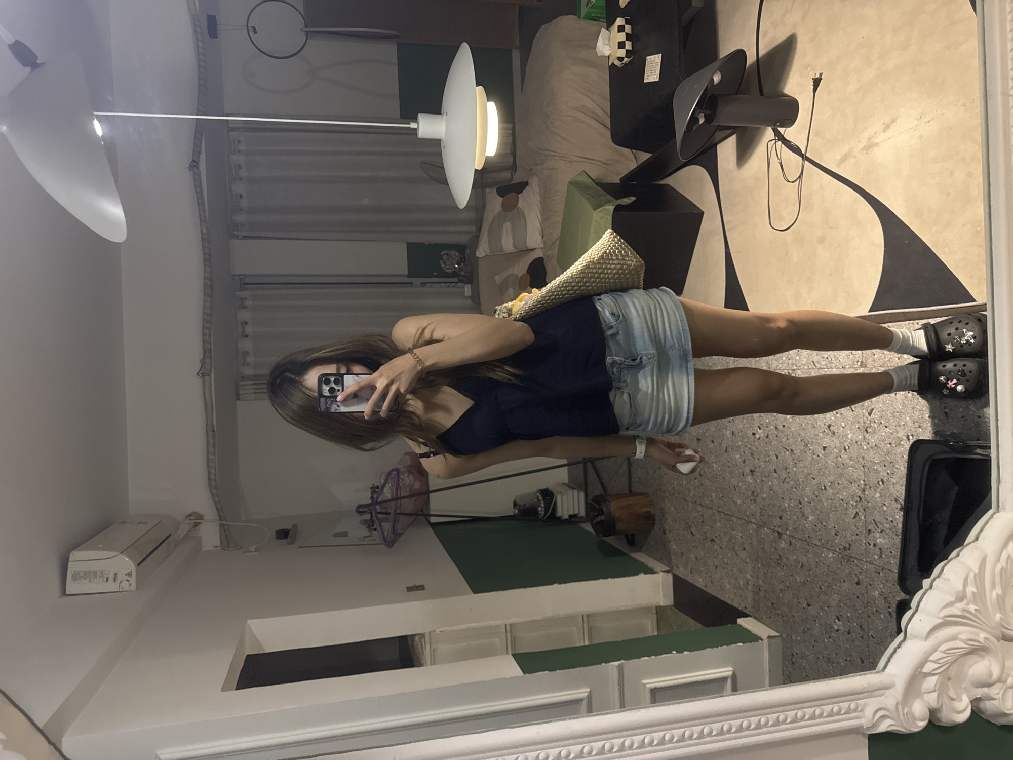 A person is captured taking a mirror selfie in an indoor setting, likely a residence in Liangjiang, China. The individual, appearing to be a young woman with long brown hair, holds a smartphone to obscure her face. She is dressed in a dark blue sleeveless top, light wash denim shorts, white socks, and black Crocs adorned with various small charms. Accessories include a white smartwatch on her left wrist, a gold-toned bracelet on her right, and a woven straw bag draped over her left shoulder.
The reflection reveals a contemporary room with diverse elements. A distinctive white pendant lamp hangs from the ceiling, illuminated. Behind the person, a bed with beige bedding and two pillows featuring abstract, wavy designs is visible. The room's walls are partly painted in a dark green accent color, contrasting with the dominant white. A black standing fan is positioned on the right side of the room, near a wall with a bold black and white abstract pattern. Various wires and cables are visible, along with a white air conditioning unit or similar device mounted on a shelf. The floor has a speckled, terrazzo-like appearance. The mirror itself features an ornate white frame along its bottom and right edges. The artificial lighting from the pendant lamp suggests the photo was taken indoors, possibly in the evening or at night. No discernible text is clearly legible on any objects within the frame.