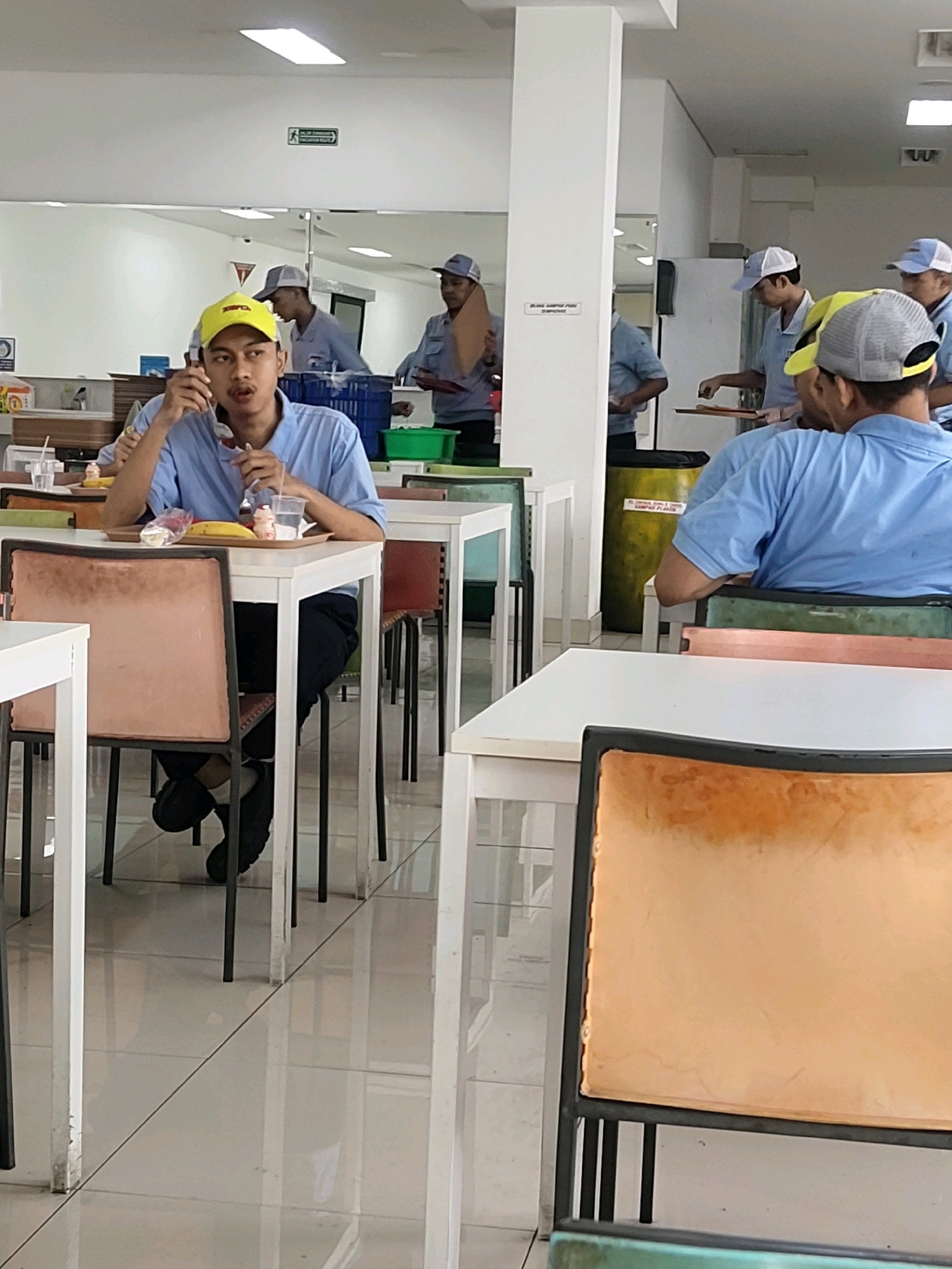 The image depicts an indoor canteen setting, likely in Mekarmukti, Indonesia, with several individuals present. In the foreground, a young man wearing a light blue polo shirt and a yellow baseball cap is seated at a white table, holding a spoon and fork, and appears to be eating from a tray. The tray contains a banana, what looks like yogurt or pudding cups, and a drink. He is seated in a chair with a padded orange seat. In the background, other individuals, also wearing light blue shirts and some with caps, are visible. Some are standing, and one appears to be interacting with a trash bin. The environment suggests a cafeteria or dining hall, with multiple tables and chairs, and a tiled floor. Reflections in a mirrored surface behind the central figure indicate additional seating and possibly a service counter area. The lighting appears to be overhead fluorescent lights.